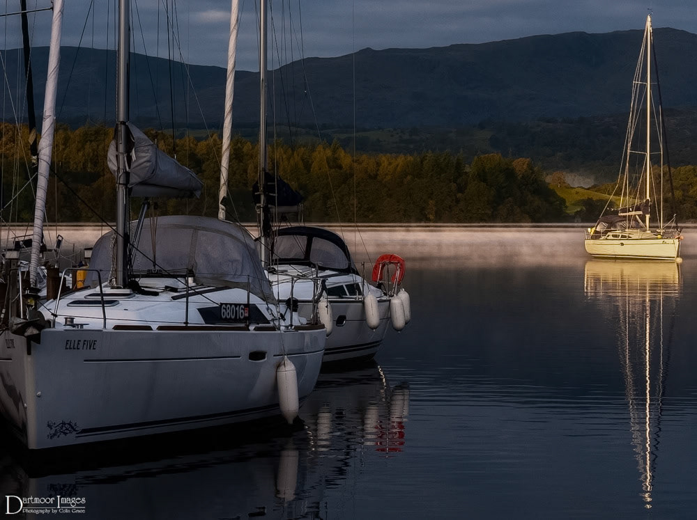 As yachts lay moored alongside the marina at White Cross Bay a slight mist lingers on the surface of Windermere in The Lake District as the early morning light falls onto a moored yacht and lights up the far shore line of the lake.