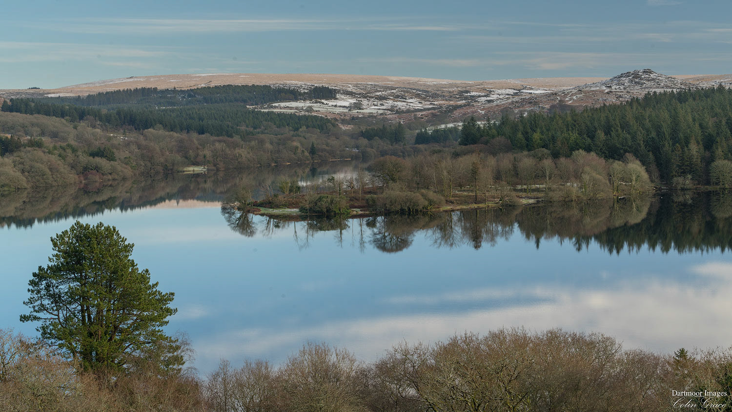 A light dusting of snow lies on Dartmoor as the trees and clouds are reflected in the still waters of Burrator Reservoir.