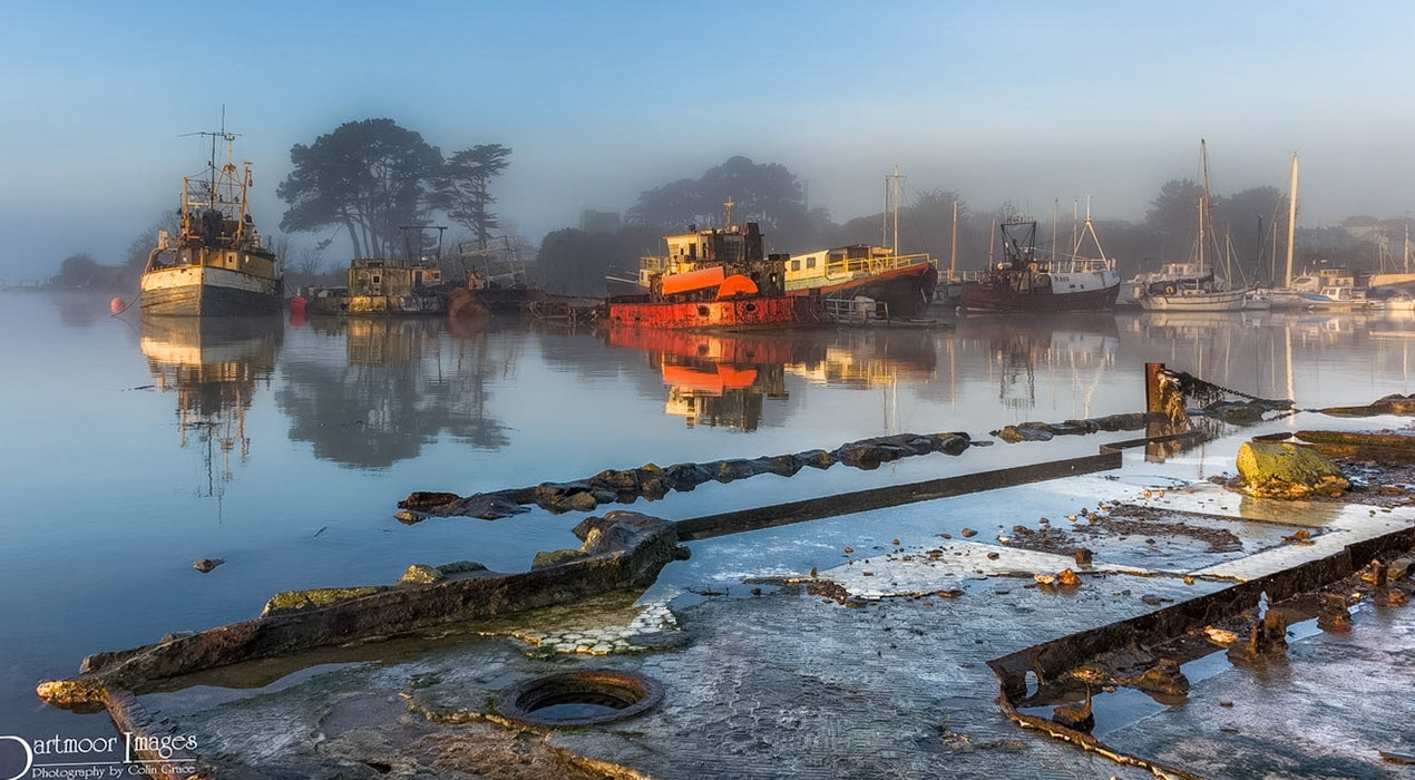 Old barges and boats moored alongside Higgins Brothers boat yard at Torpoint in Cornwall during a misty sunrise.