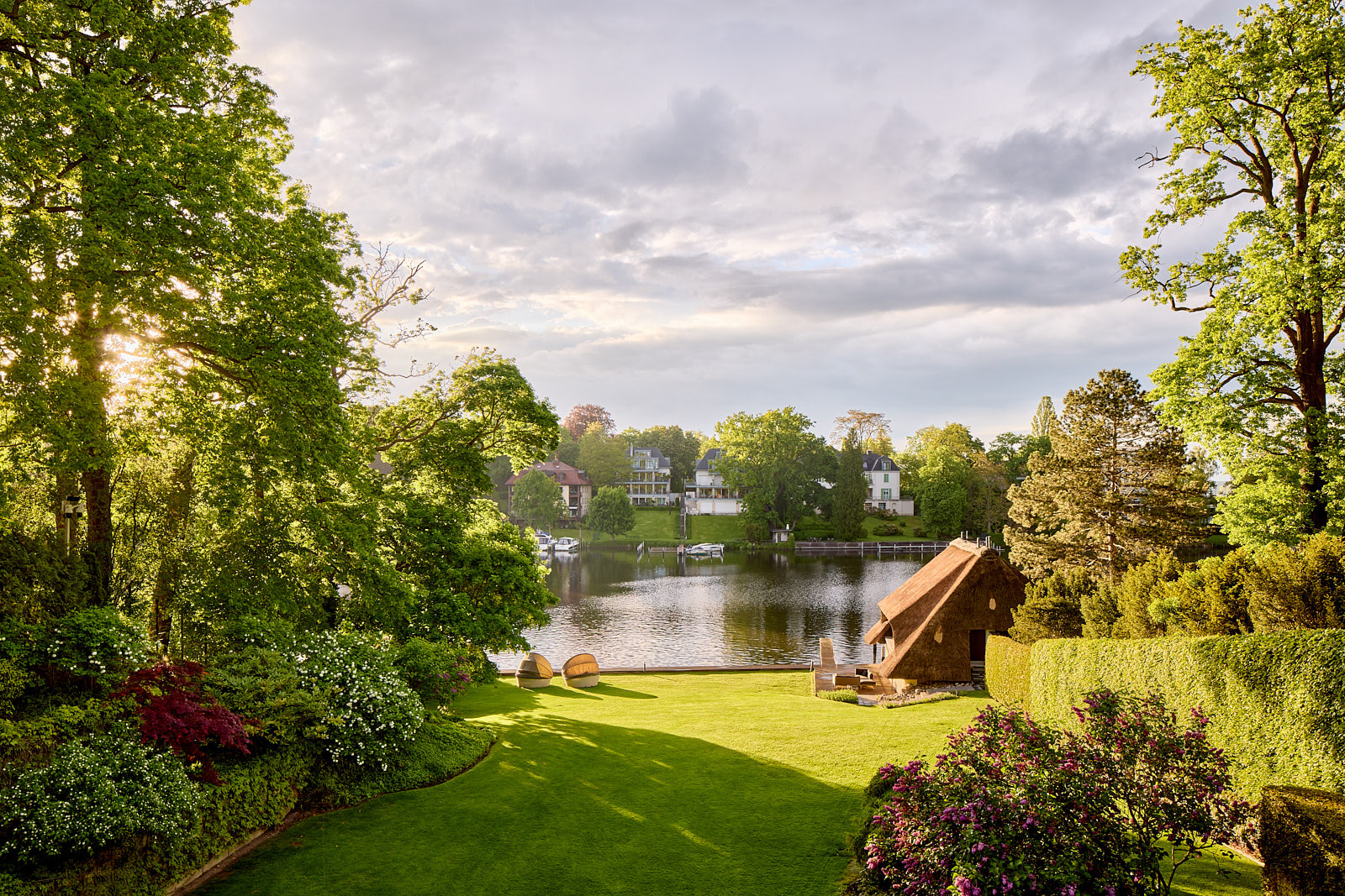 Parklandschaft eines privaten Anwesens mit Wasserfläche, offenen Rasenflächen und dichtem Baumbestand.