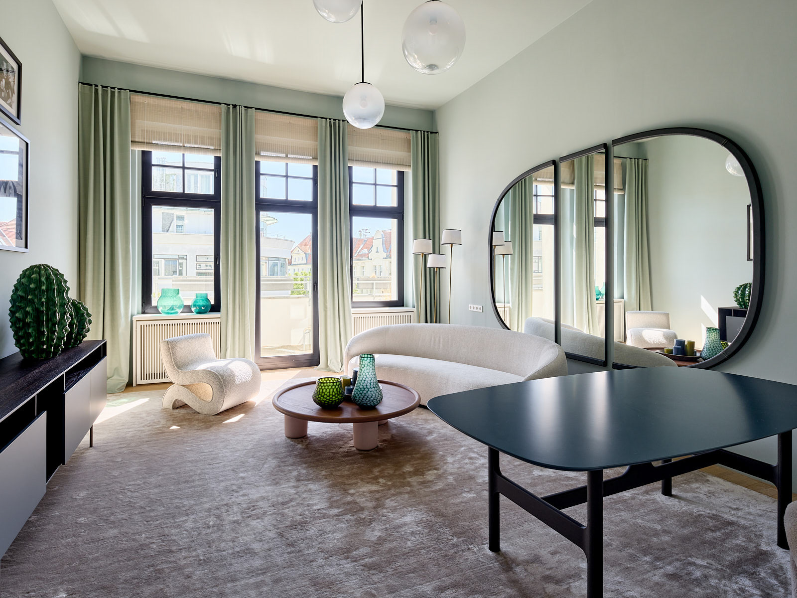 Hellgrüner Salon mit geschwungenem hellem Sofa, großem mehrteiligem Wandspiegel und schwarzen Fensterrahmen.