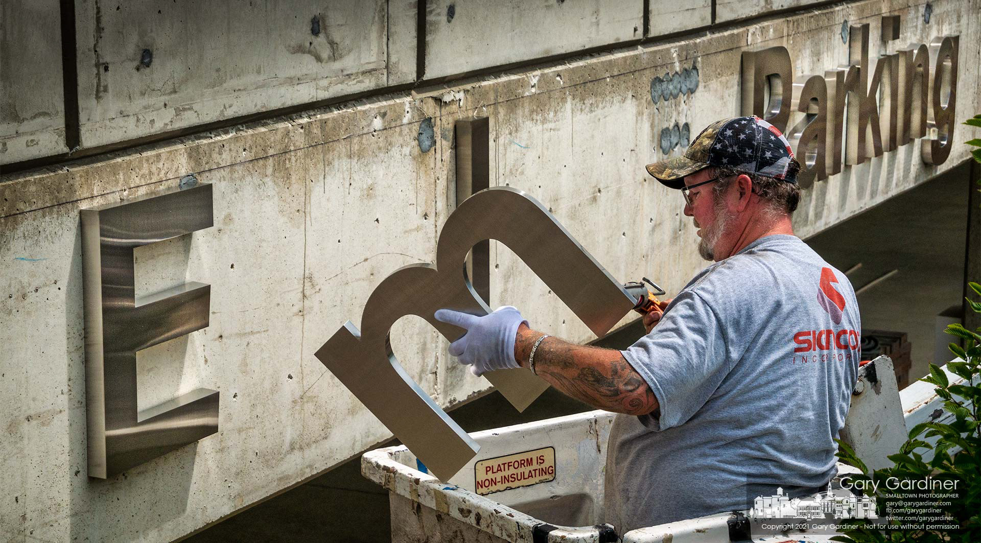 A signmaker applies adhesive to the letter "m" before attaching it next to the capital "E" to create signage for the employee parking entrance at the COPC building on Africa Road. My Final Photo for July 16, 2021.