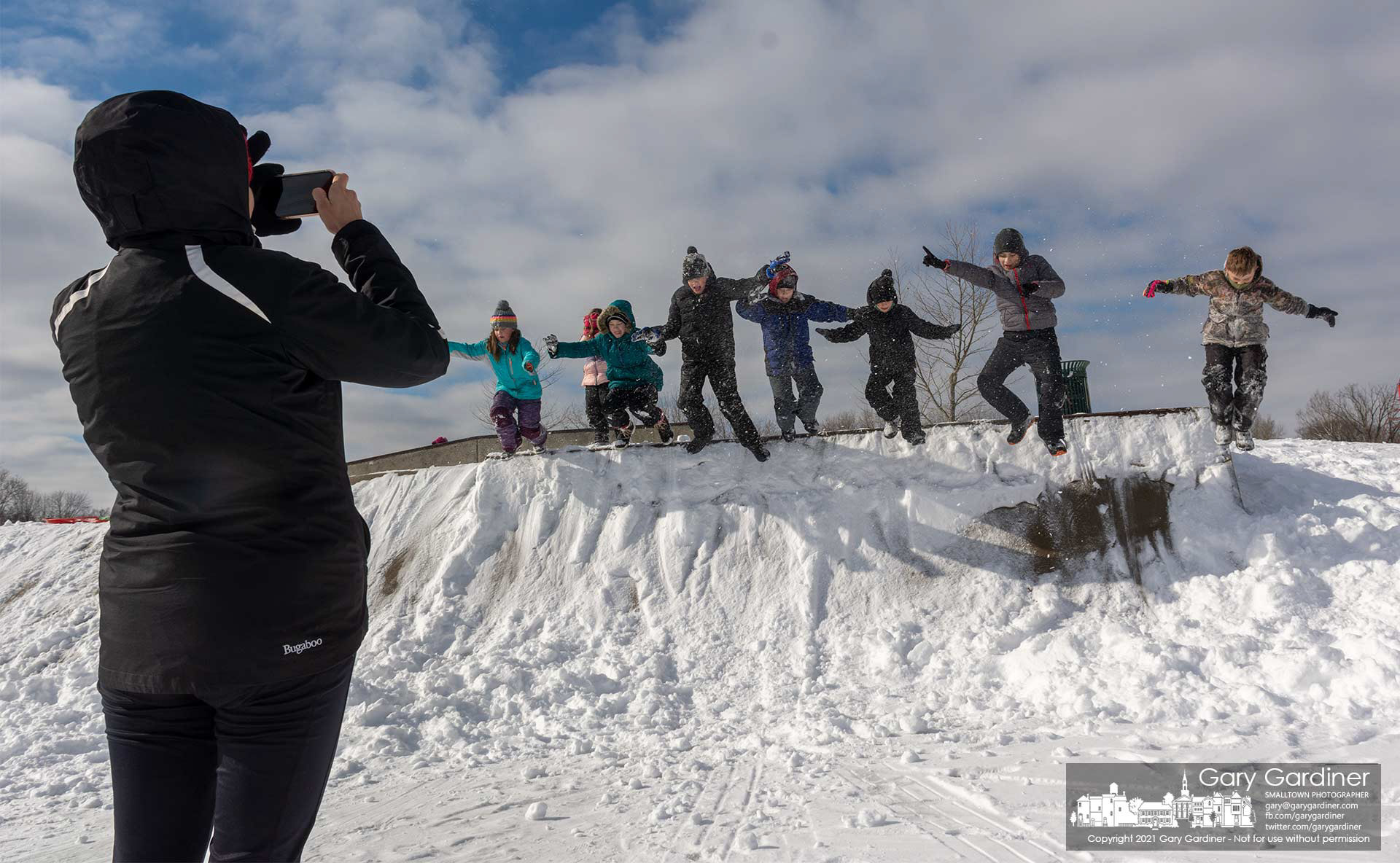 A mother photographs her children and their friends as they leap off the lip of the quarter-pipe section of the Westerville Skate Park where they chose to sled the shorter hill next to the newer sledding hill. My Final Photo for Feb. 2, 2021.
