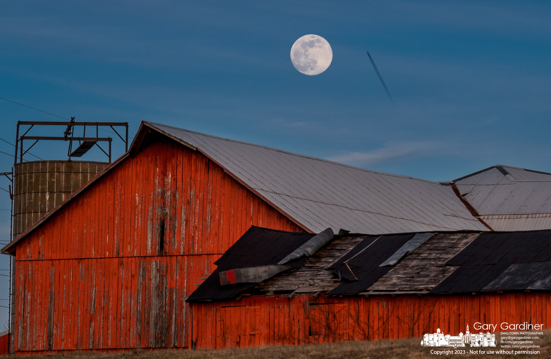 A jet leaves a contrail across the sky as the February full moon rises behind the Yarnell Farm barn. My Final Photo for February 4, 2023. 