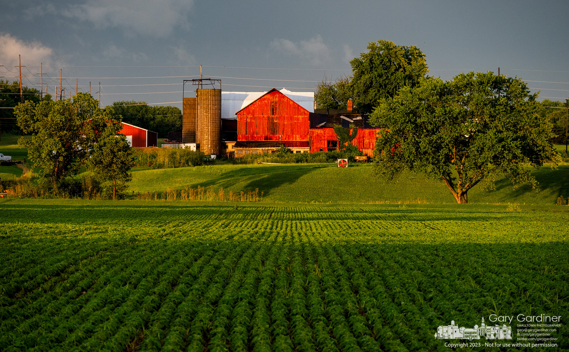 The setting sun casts shadows across a soybean field below the Yarnell Farm barns that are bathed in lights moments before the sun disappears below the horizon. My Final Photo for July 2, 2023. https://bit.ly/46uUbB7