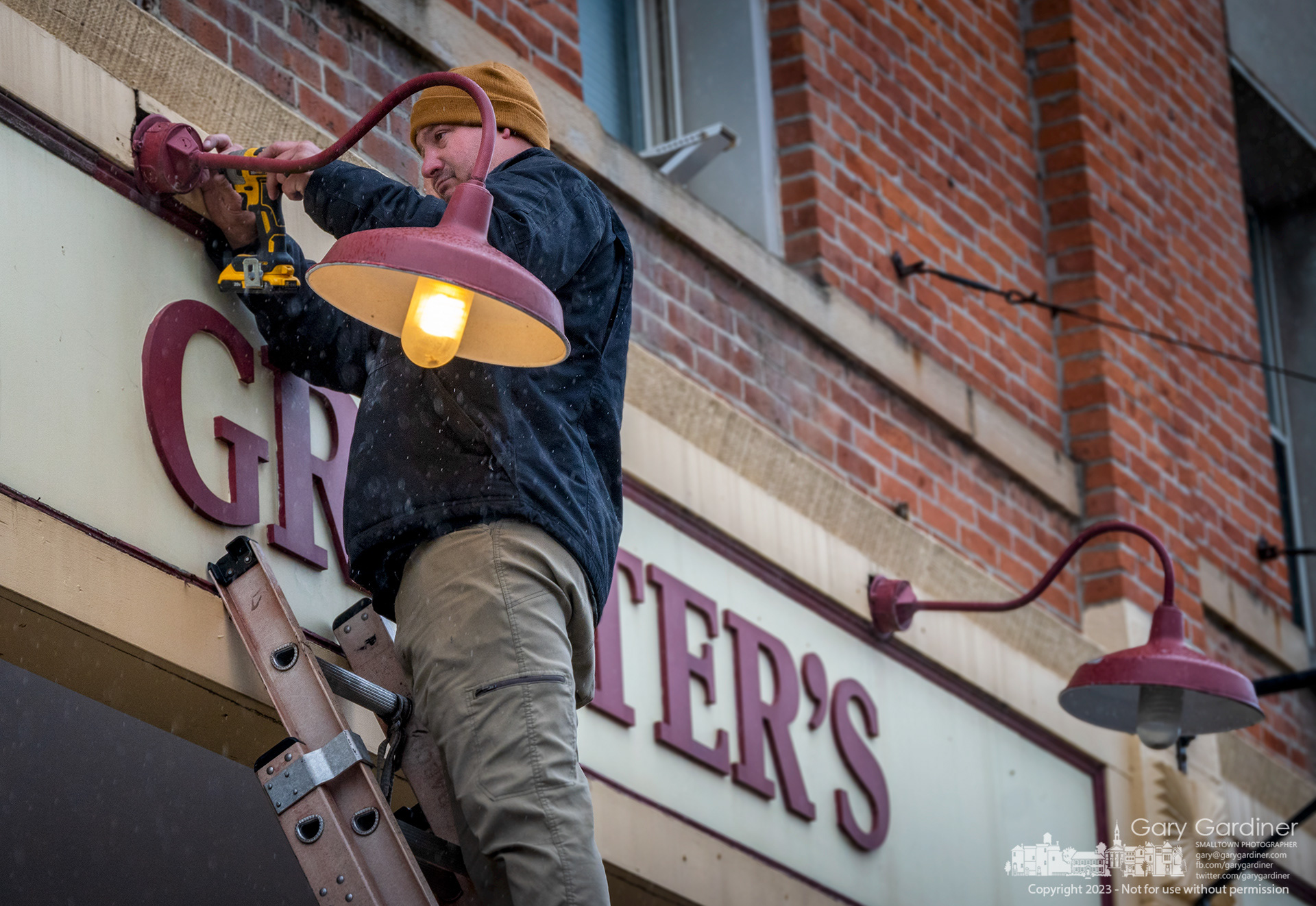 An electrician anchors a repaired light fixture to the Graeter's sign in front of the Uptown Westerville ice cream store. My Final Photo for May 2023. Photo Copyright 2023 Gary Gardiner. Not to be used without written permission detailing exact usage. Photos from Gary Gardiner, may not be redistributed, resold, or displayed by any publication or person without written permission. Photo is copyright Gary Gardiner who owns all usage rights to the image.