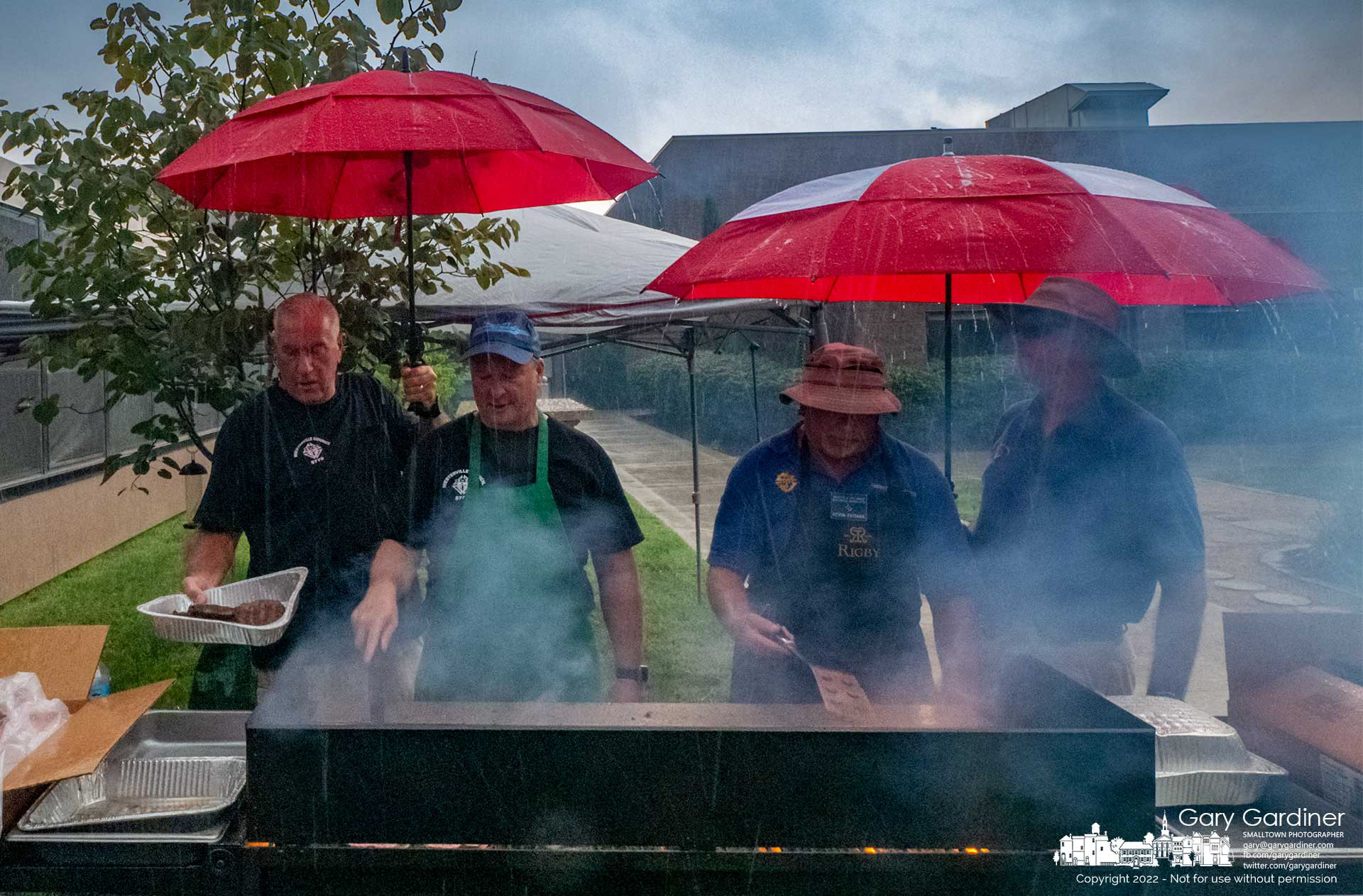 The grill cooks stand under umbrellas to shield themselves and the hamburgers and hotdogs they are preparing for the St. Paul Parish Picnic which moved indoors to stay out of the rain. My Final Photo for August 21, 2022,
