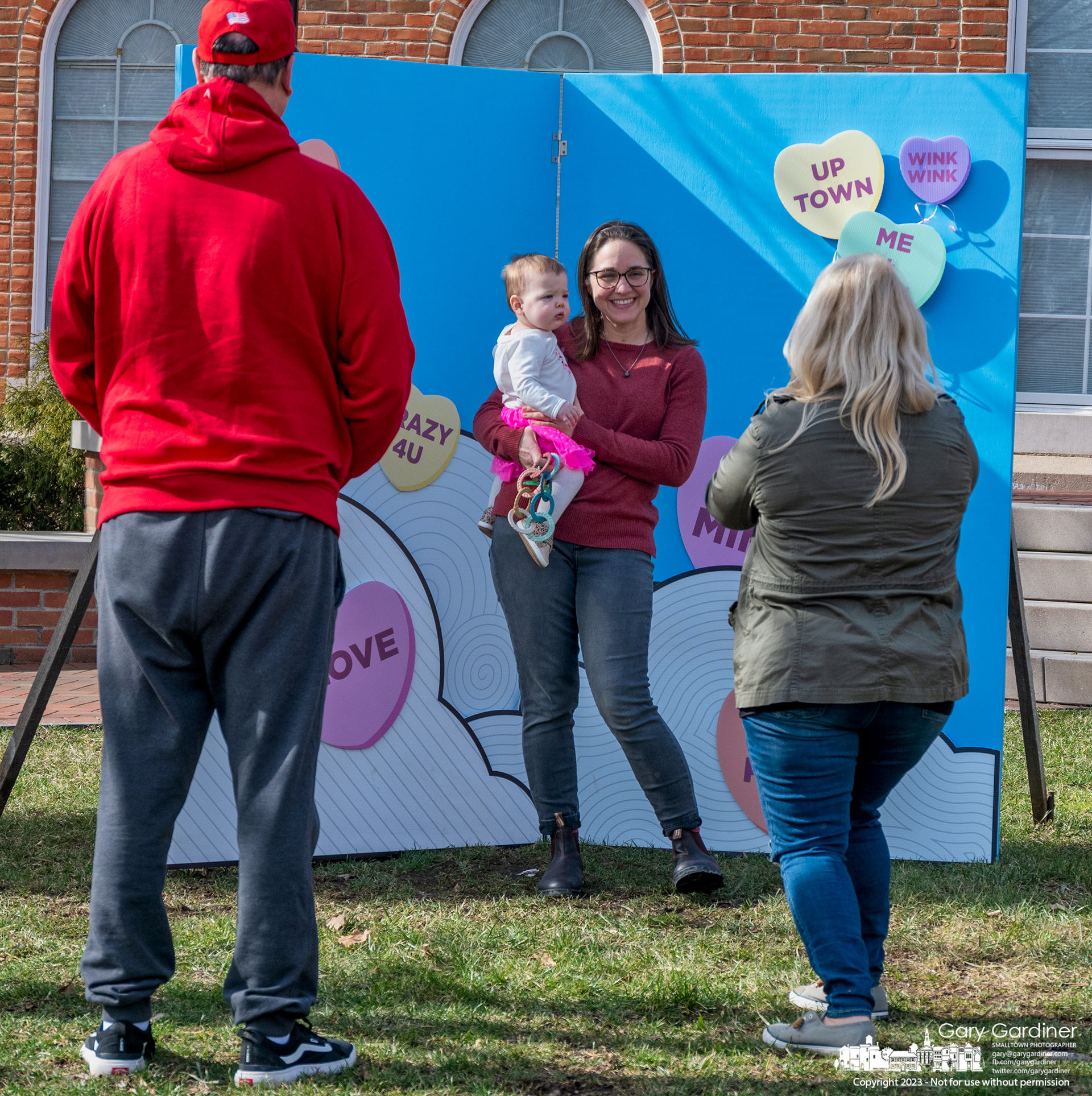 A woman and her youngest child post in front of a Valentine's Day photo backdrop on the green at city hall as a couple makes their photo in return for the woman previously photographing them in front of the banner with heart wishes. My Final Photo for February 14, 2022.