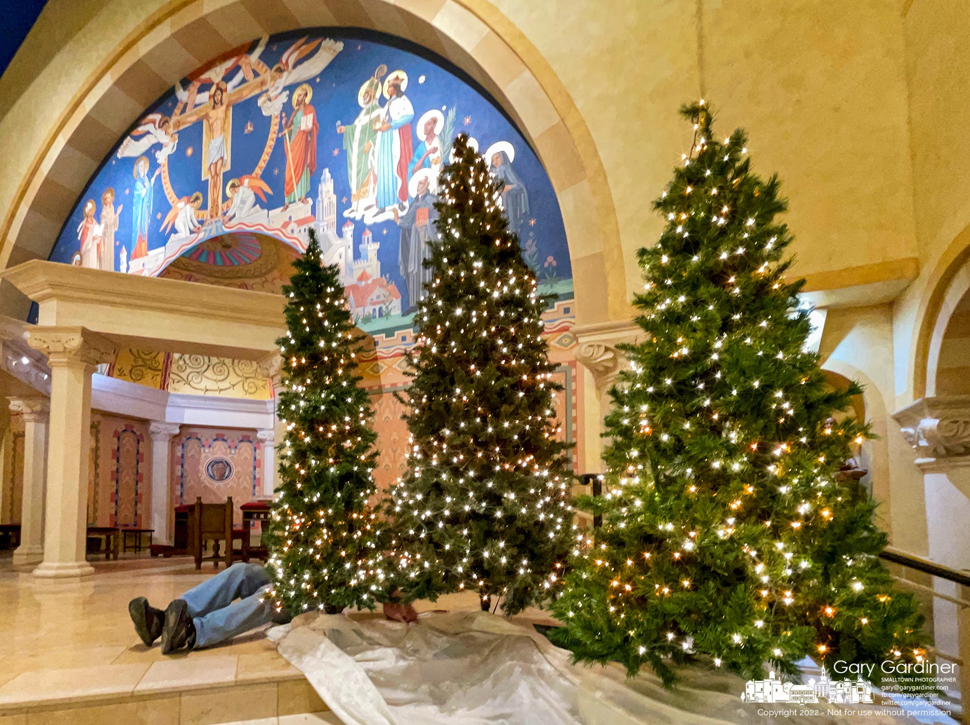 A volunteer lies beneath a Christmas tree with his legs hanging out as he places three Christmas trees near the altar preparing St. Paul the Apostle Catholic Church for Christmas day services. My Final Photo for December 22, 2022.