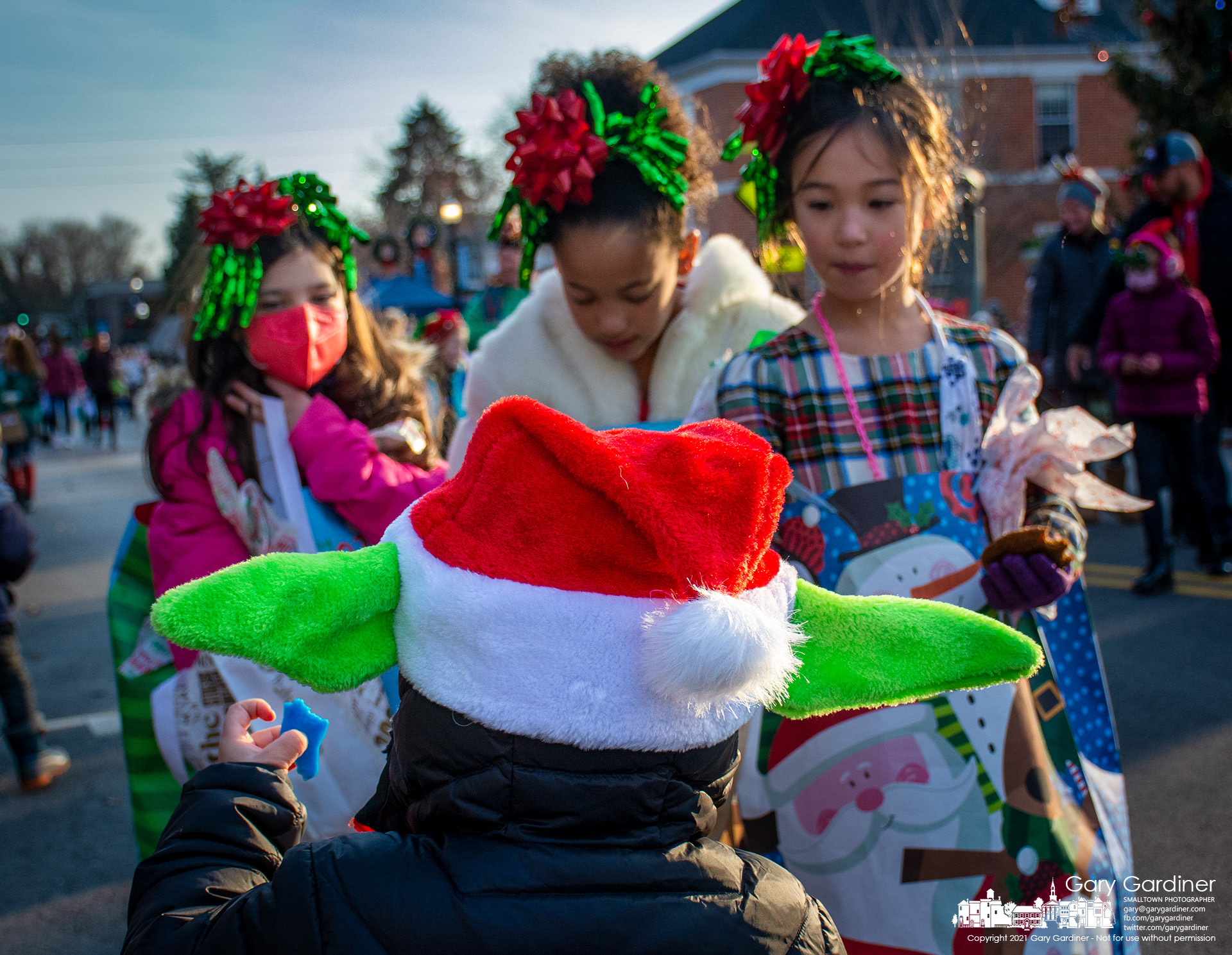 A youngster wearing a festive Baby Yoda Christmas hat with one of his hands holding a blue piece of candy is offered more candy by three girls forced by his unique holiday cap to attend to his sugary request. My Final Photo for Dec. 5, 2021,