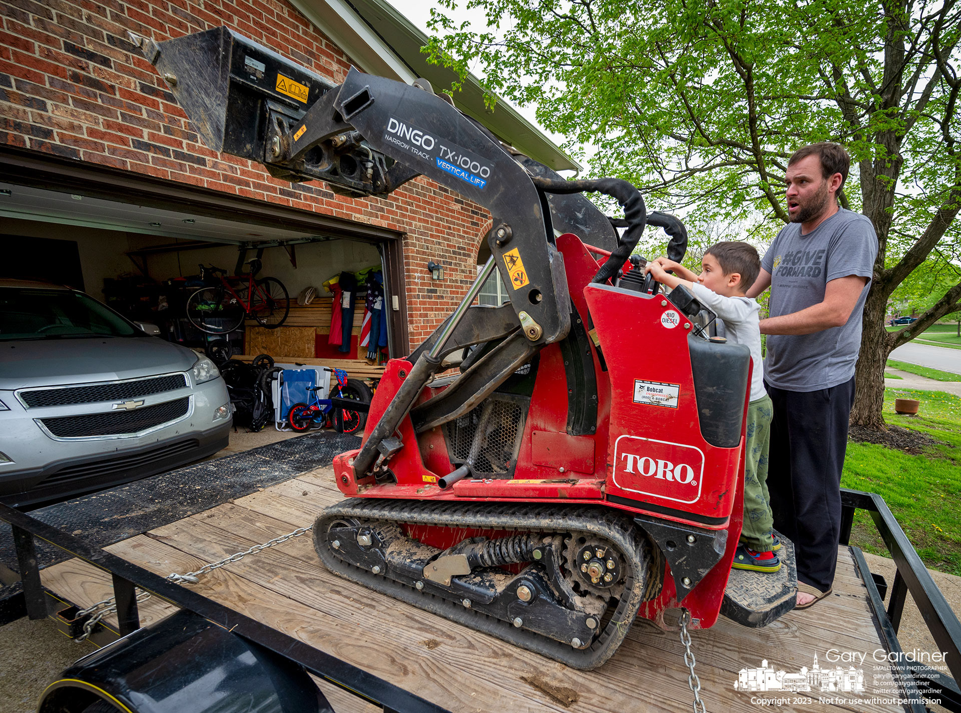A father shows his son and a few neighborhood kids how the front-end loader he uses at work allowing them to raise and lower the bucket while it is anchored to the trailer. My Final Photo for April 28, 2023. 