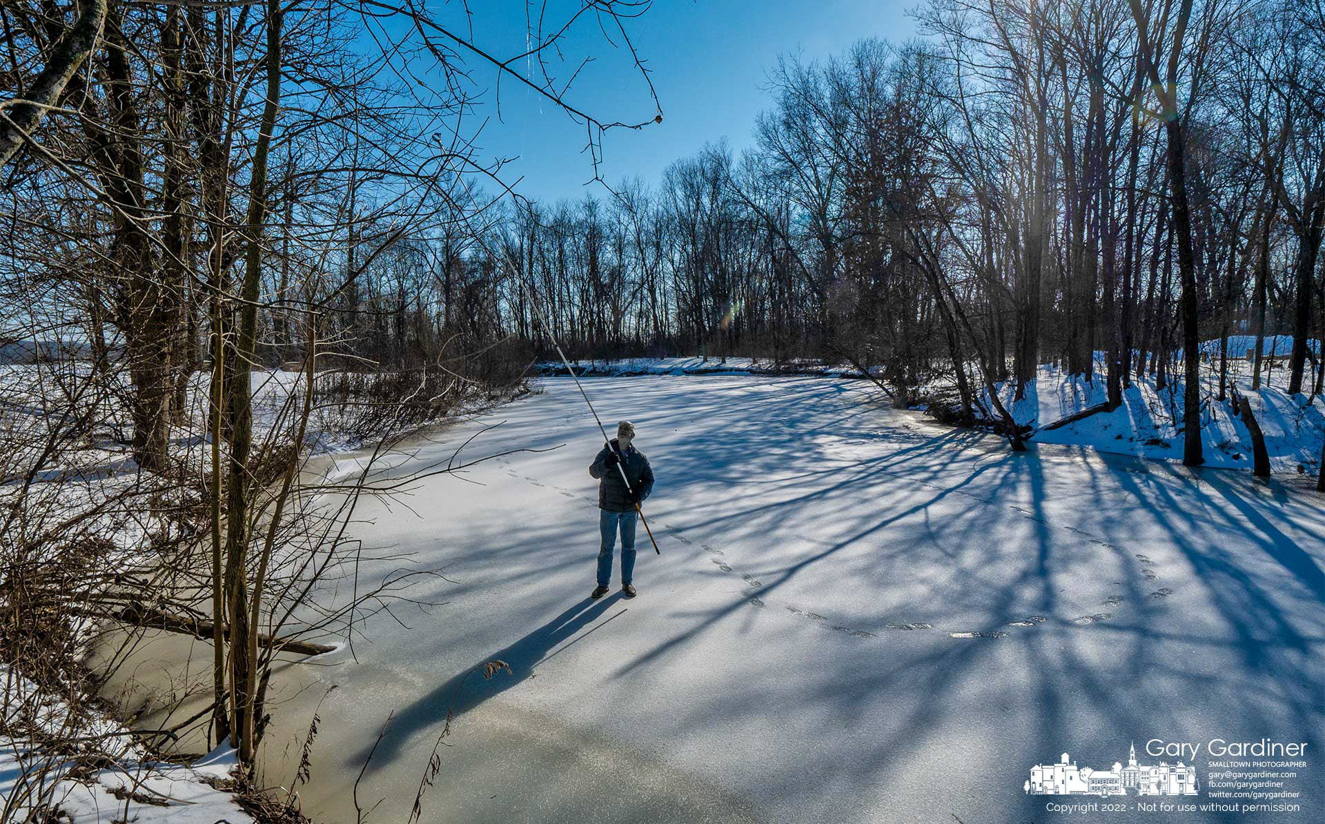 A  winter angler stands on frozen Hoover Reservoir using a telescoping cutter to retrieve other angler's lures caught in trees that hang over the water and are difficult to grab when the lake is not frozen. My Final Photo for Feb. 8, 2022.