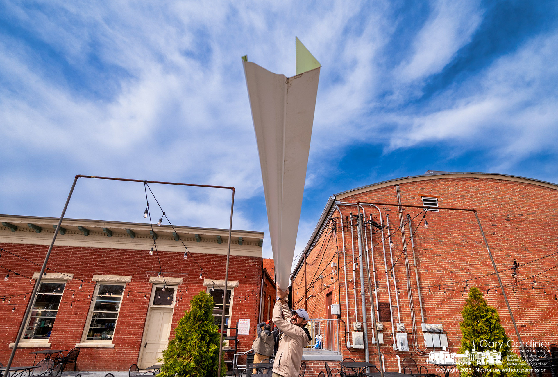 Three workers transport a 30-foot-long section of rain gutter across a parking lot toward its installation on the roof of a building in Uptown Westerville. My Final Photo for April 7, 2023.