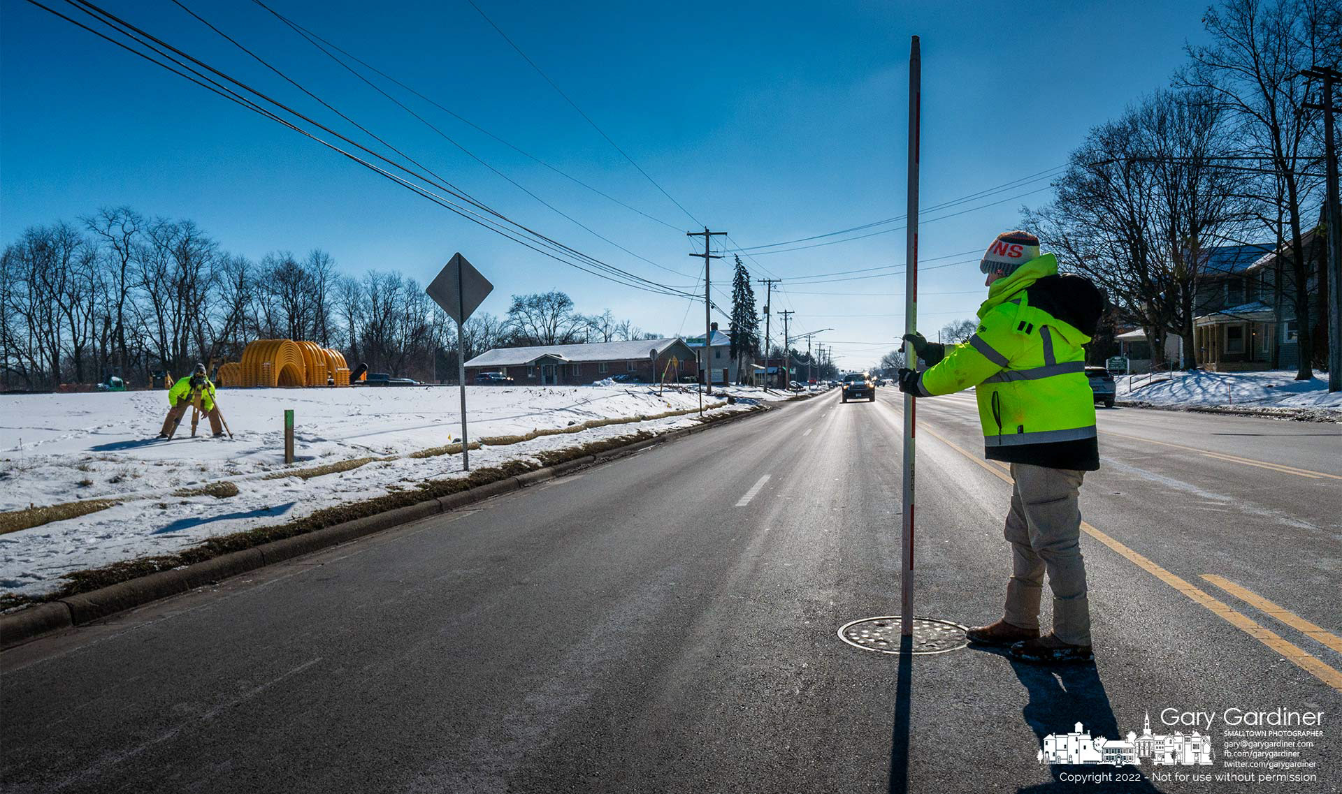 A surveyor stands over a manhole cover in South State Street where a second surveyor measures elevations for construction to begin on an office building with apartments adjacent to the post office. My Final Photo for Jan. 26, 2022.