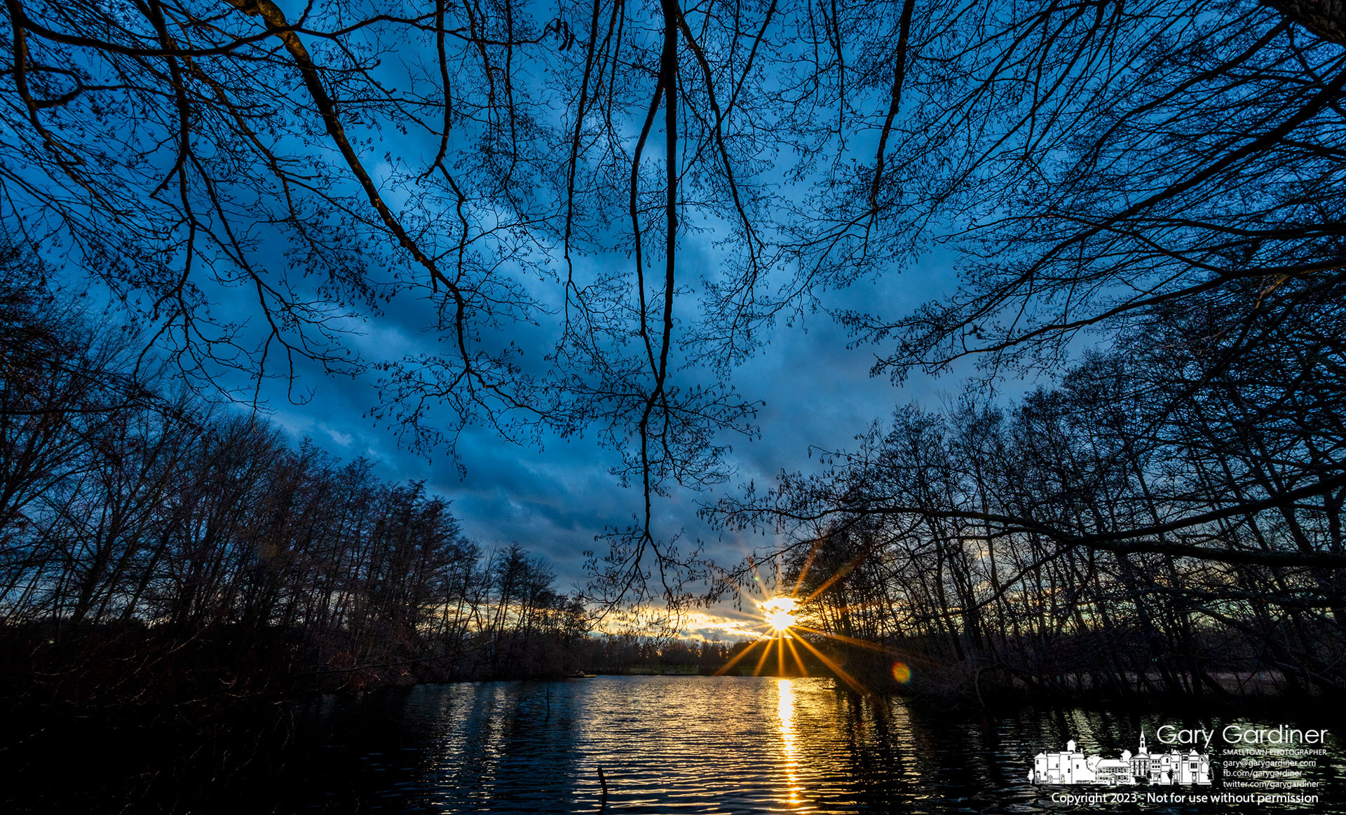 Barren branches hang over Schrock Lake at Sharon Woods Park where the sunset is reflected in the slightly rippled waters of the lake. My Final Photo for January 17, 2023.