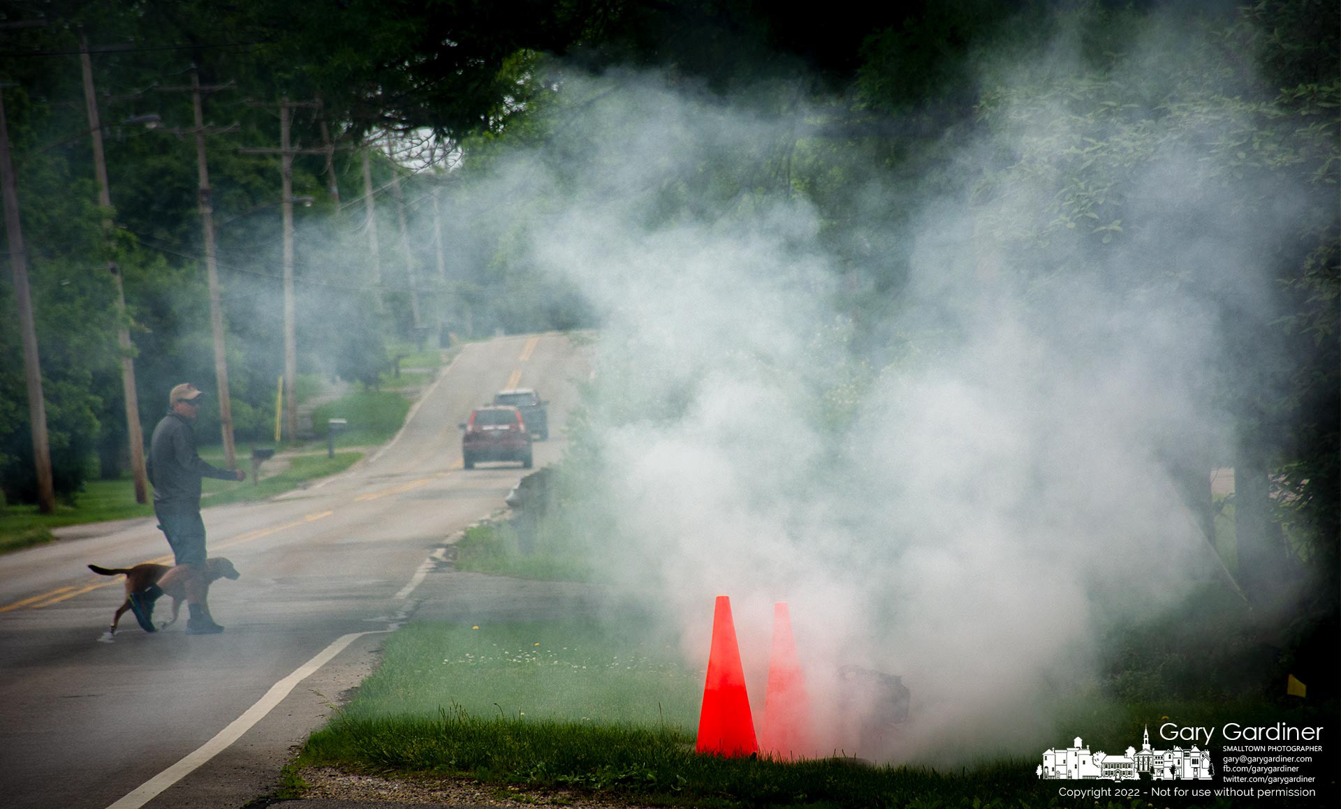 A man and his dog cross East Walnut street near an open manhole on a sanitary sewer spewing smoke used for testing the integrity of the sewer system. My Final Photo for May 23, 2022.