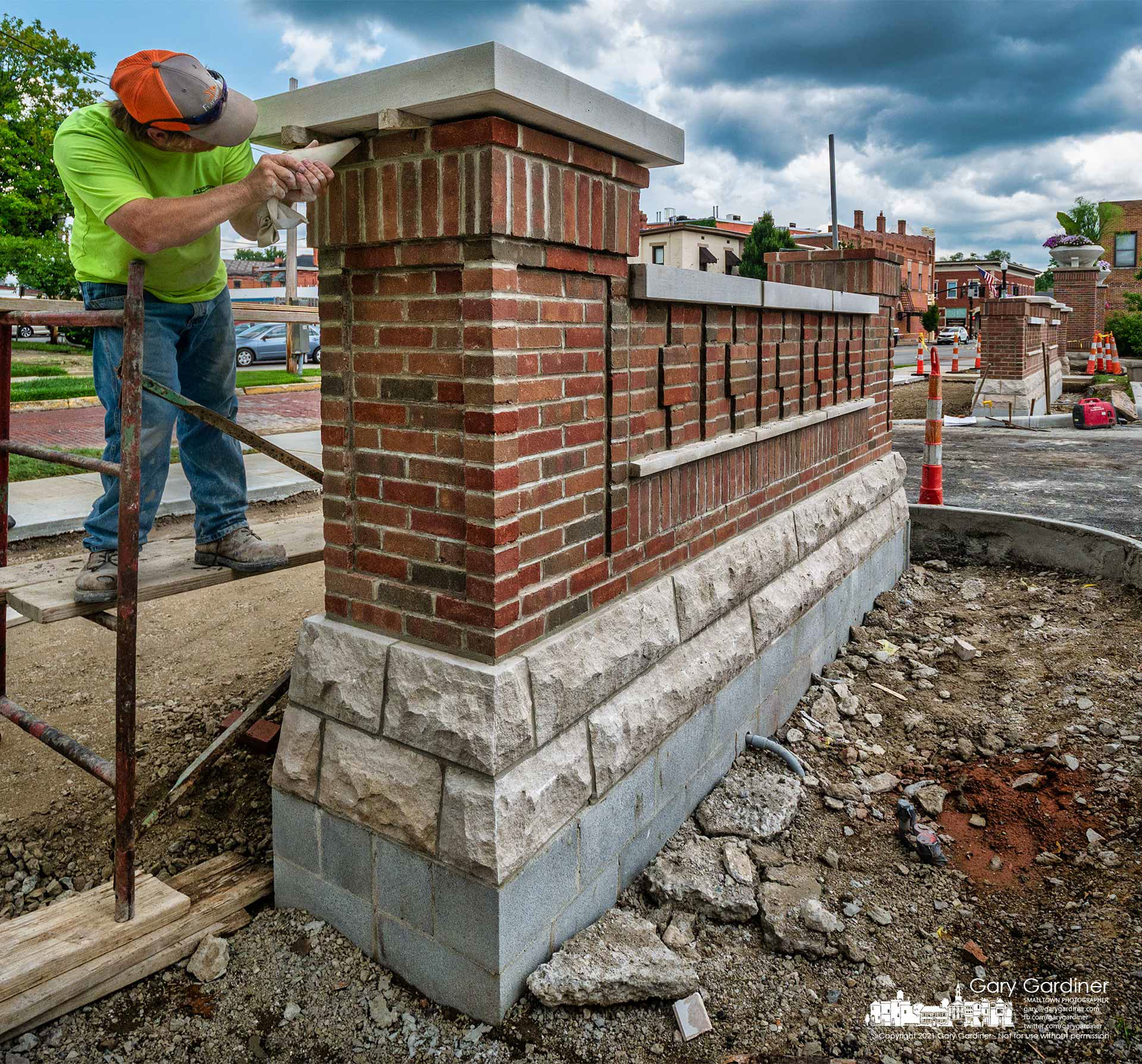 A brick mason squeezes fast-drying mortar into the gaps between the brick post and a concrete platform lowered onto it at the entrance to the new parking lot behind city hall. My Final Photo for July 14, 2021. 