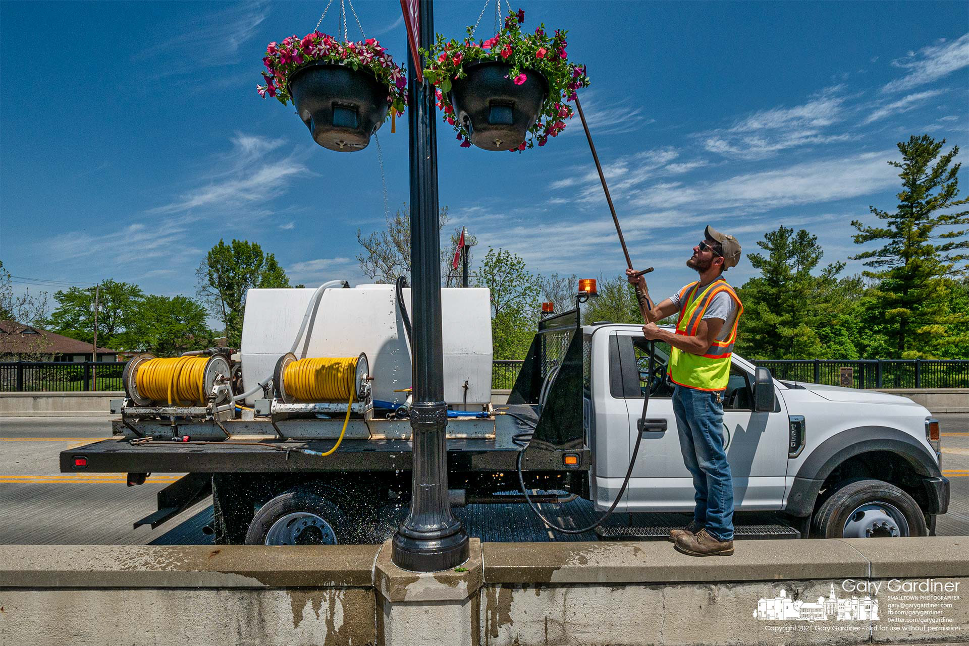 A city parks worker stands on the concrete barrier as he irrigates hanging baskets on the Main Street Bridge. My Final Photo for May 18, 2021.
