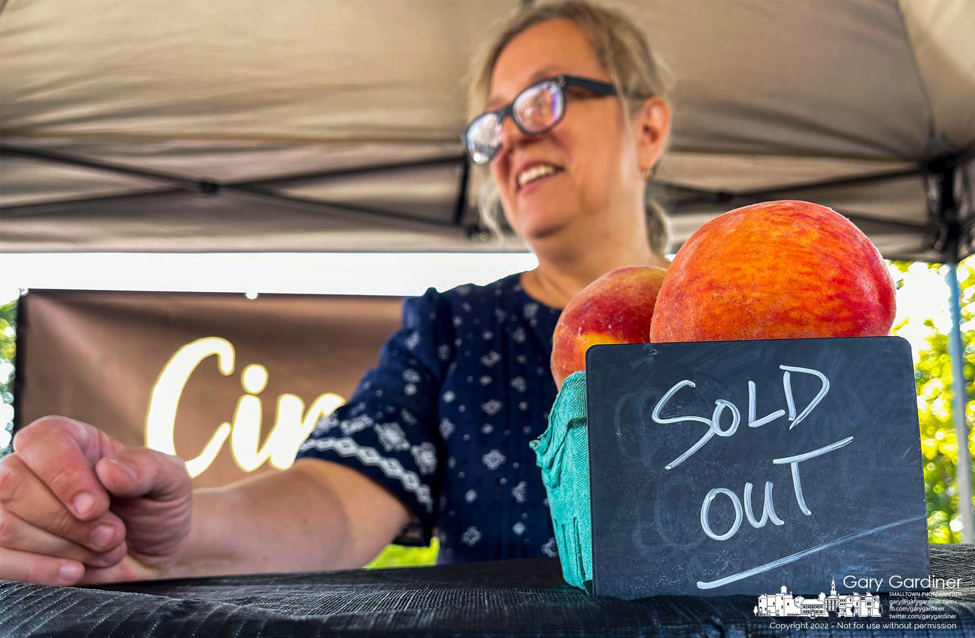 A cinnamon bun baker uses a neighbor vendor's fruit and display box to hold up her "Sold Out" chalkboard less than halfway through the Saturday Farmer's Market. Several vendors sold out early with the large crowd arriving early in the morning. My Final Photo for August 27, 2022.