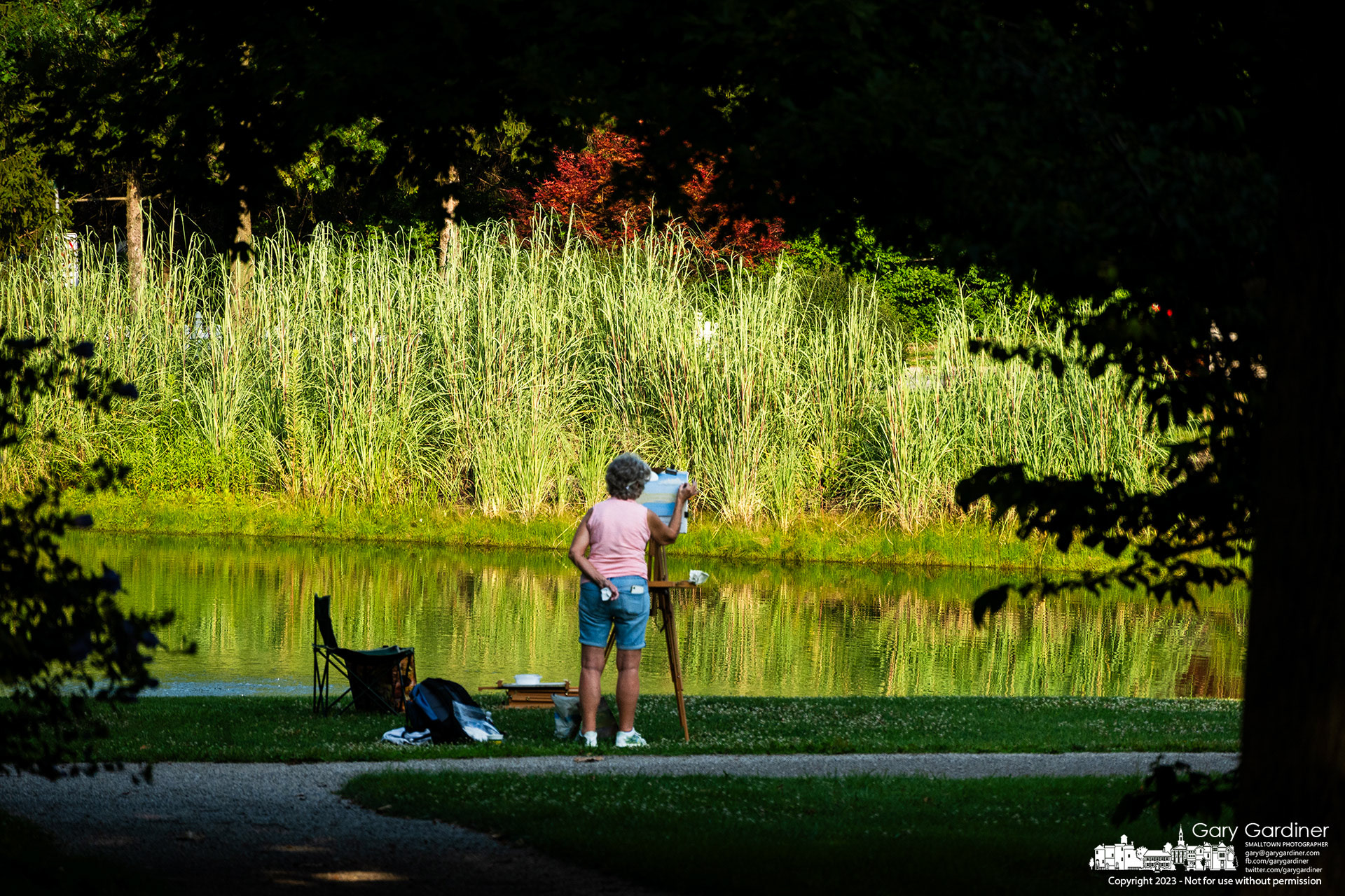A plein air artist applies chalk pastels to her canvas as she stands in the late afternoon shadows edging over the pond at Heritage Park. My final Photo for August 3, 2023. https://bit.ly/3DKiTAp