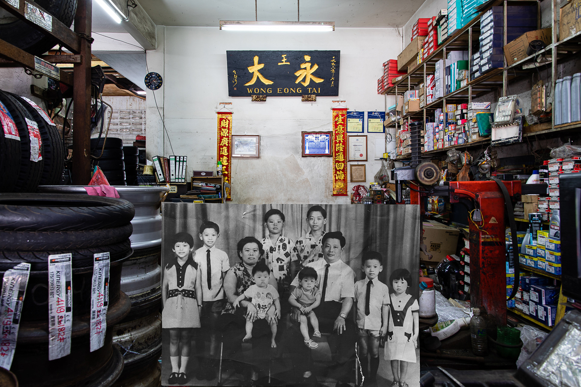 黑白图像为1971年,祖父母与叔叔、姑姑们在影楼拍摄的第二张全家福。图片背景为当今的店面。 The superimposed B&W image was the second family portrait consisting of my grandparents, uncles and aunts taken in a photo studio in 1971. The background photo showed the current shop front of the family-owned tire shop.