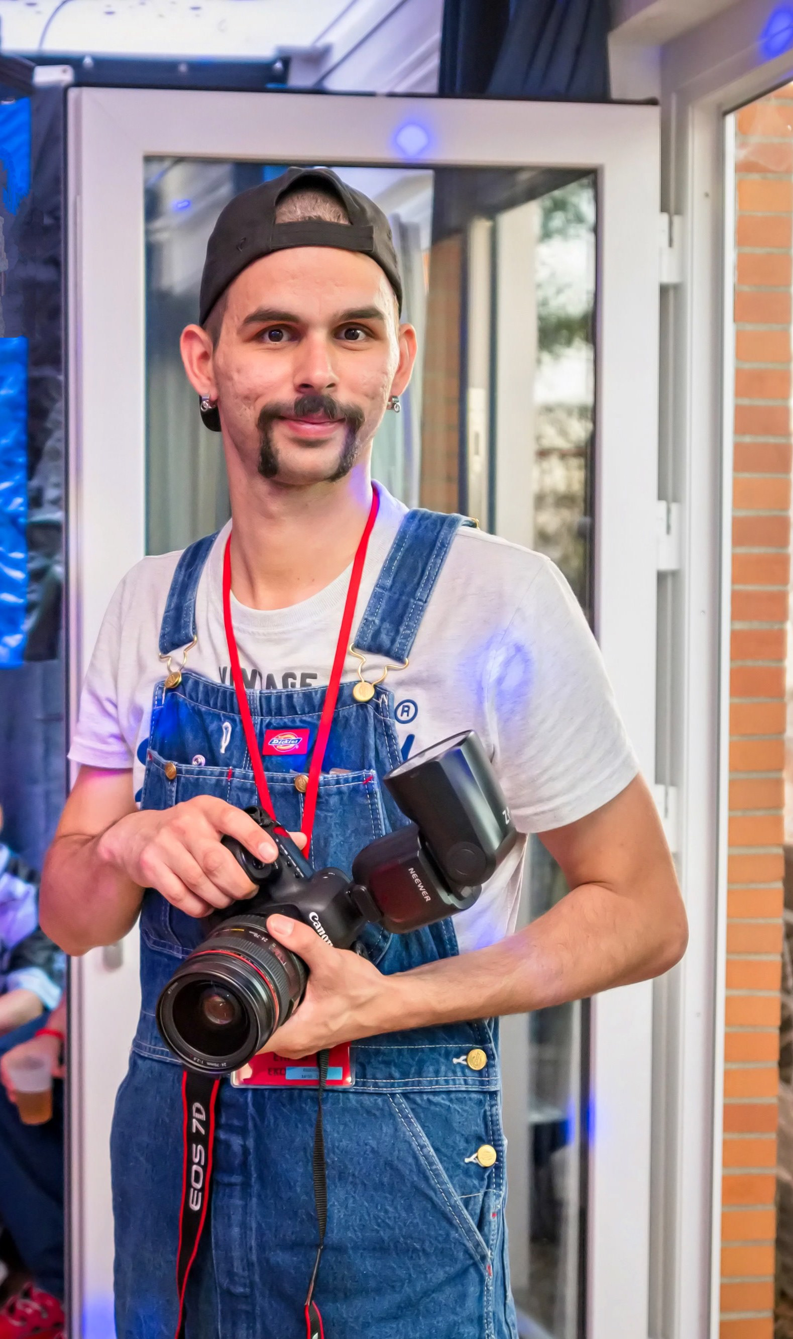 Enrico Sanchez, mit Mütze und Latzhose, mit seiner Kamera Canon EOS 5D Mark IV und Blitzgerät bei einer privaten Veranstaltung. EKOSphotography