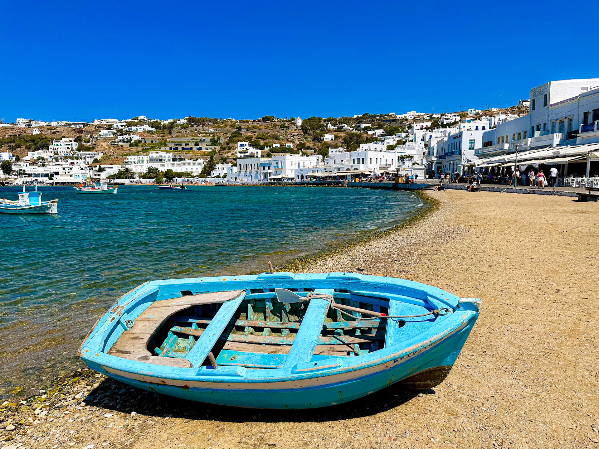 Azure Anchorage: Mykonos Moments: In the heart of Mykonos, an old blue rowboat rests ashore, juxtaposed against the cerulean ocean. Beyond, the iconic white washed buildings of Mykonos city stand as a silent witness to timeless Greek charm.