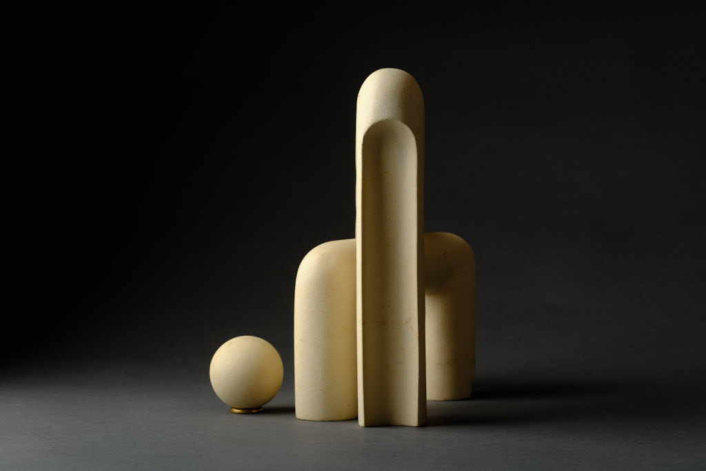 A maltese limestone tubular sculpture with three elements. A larger, bridging element with one vertical taller than the other crosses over a shorter bridging element with equal supports. A seperate sphere is balanced on a bronze ring. Arched elements are removed from the supporting elements. The sculpture is maltese limestone and photographed against a dark grey background. 