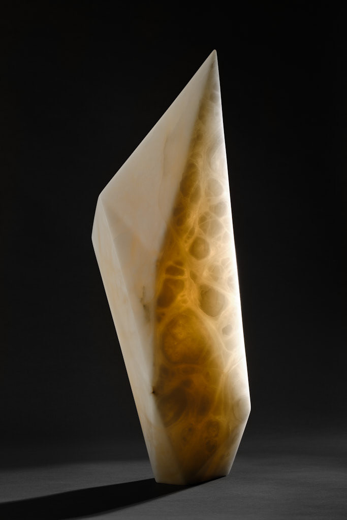 A white alabaster 'shard' sculpture against a dark background. A light source is behind the sculpture highlighting the inclusions in the stone. The sculpture is tall, narrow and angular with a directional empahasis. 