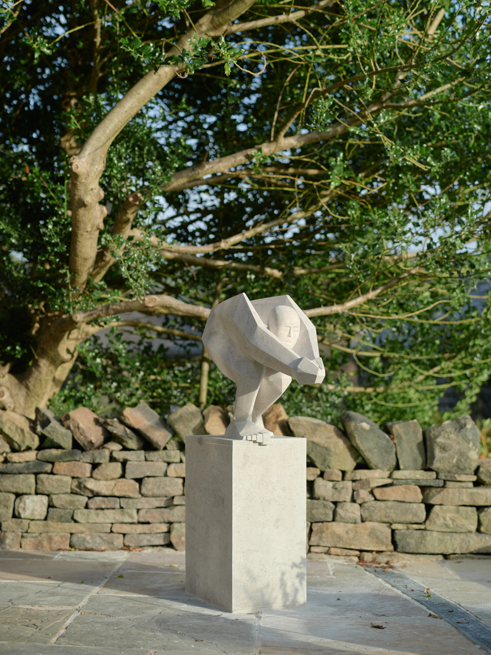 A photograph of a light grey stone sculpture installed in a garden. The sculpture is a female figure carved in stone in an abstract/ expressionistic form. The form is poised to dive from a plinth.The hands are clasped in an arrow formation. The shoulders and legs are emphasised. There is a holly tree behind, dry stone walls and paving.