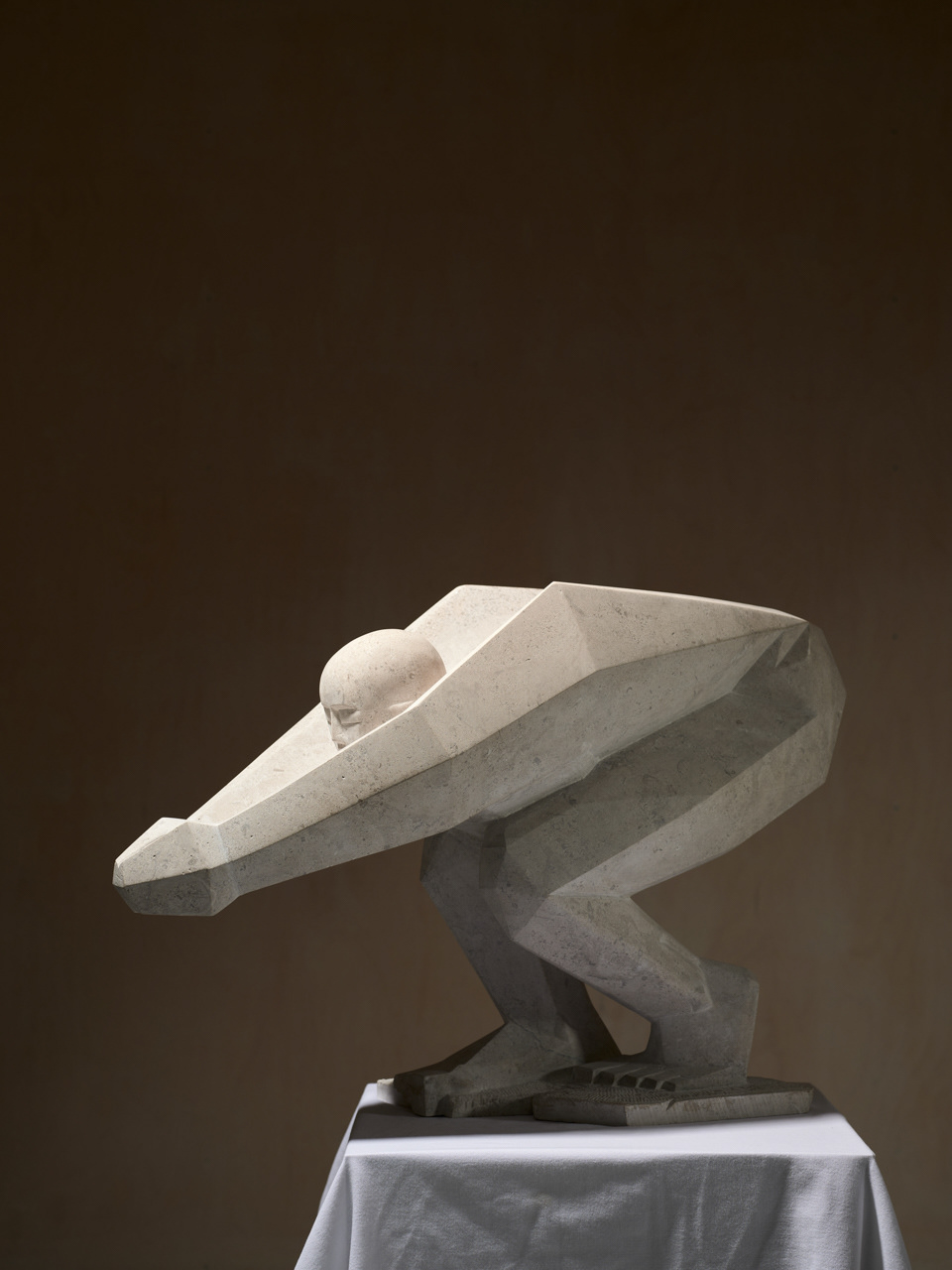 Photograph of a stone sculpture of a 'Diving Form' standing on a temporary plinth. The form is an angular interpretation of a female figure with emphasis on the strength of the legs and arms. The light grey stone sculpture is photographed against a dark background. Shadows accentuate the facets of the form.