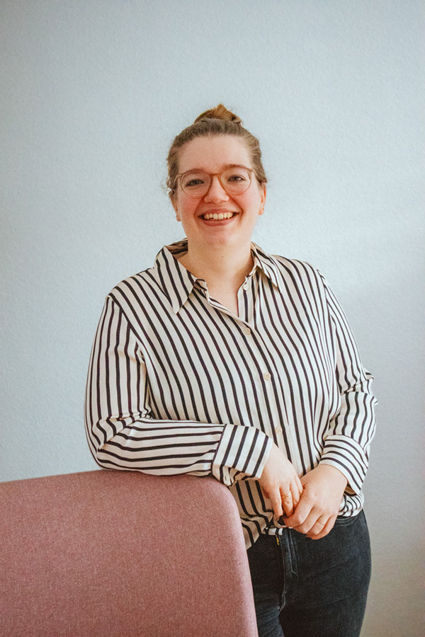 Eine Frau mit Brille in einem gestreiften Hemd steht entspannt an einem rosa Stuhl.