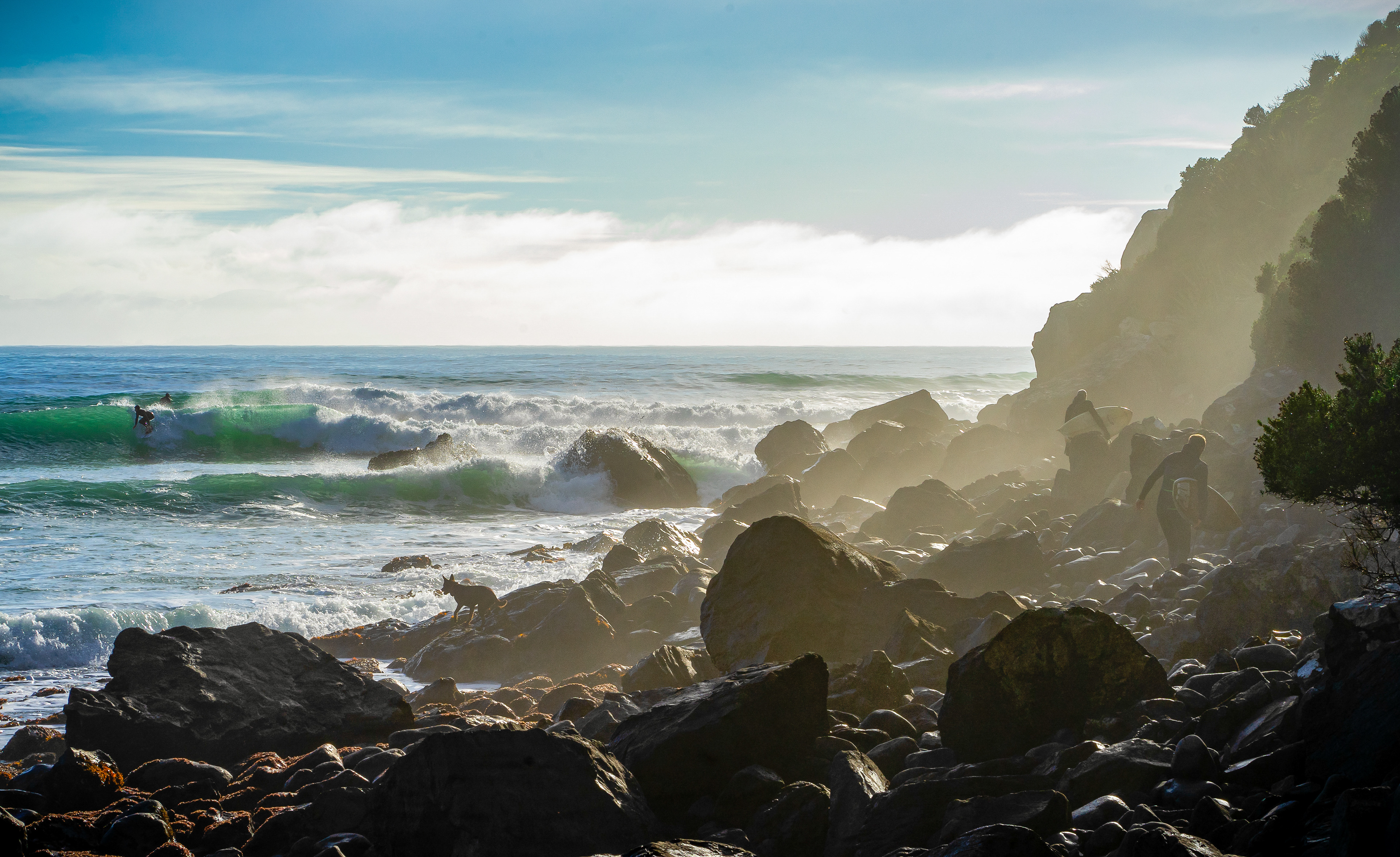 Two surfers walk over rocks on the shore while others surf the waves off the cost of Dunedin on the South Island of New Zealand on June 14, 2018.