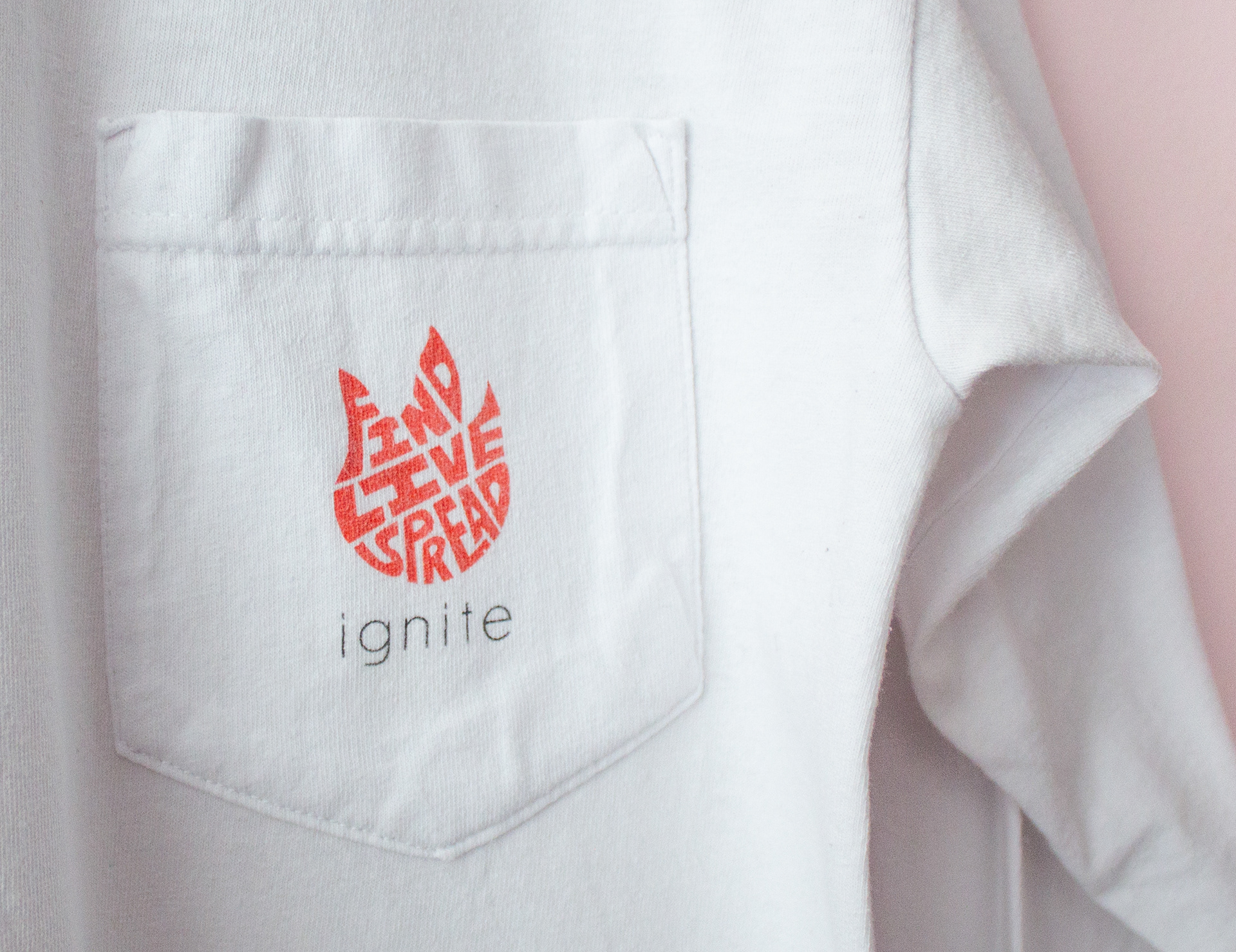 A closeup of a white long sleeve pocket shirt with an orange flame logo made of the words "FIND", "LIVE", "SPREAD". Below the logo is the word "ignite" in thin black letters.