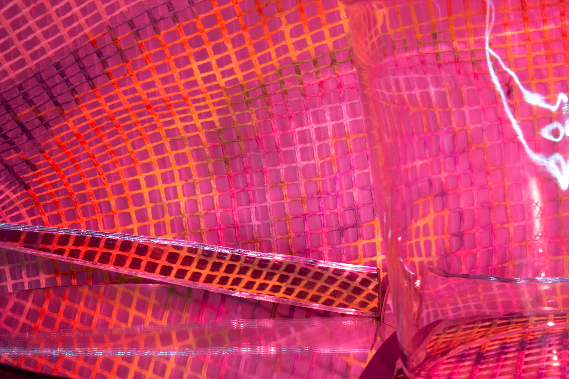 This is an abstract photograph that uses a glass and multiple mirrors to create confusion and curiosity. Overall, the image is very pink. In the background there is a net of some sort. This net has rings of different color on it. Below the multicolored net are two mirrors that reflect the lines of the net. On the right is glass that distorts the lines of the net behind it. It also creates a bright white glare.