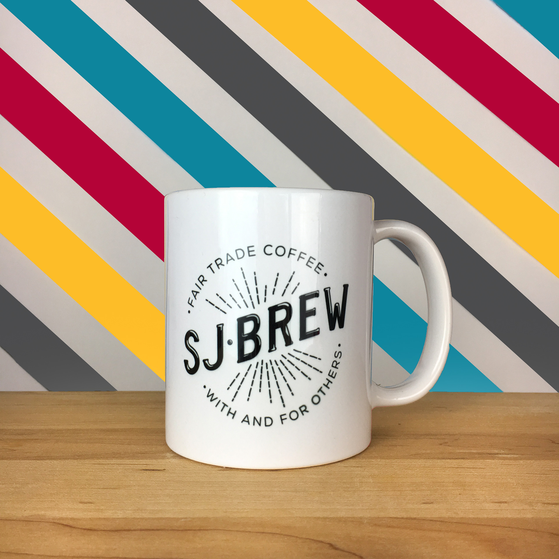 This square ratio photo depicts a white coffee mug with an SJ Brew logo in black. The logo reads “FAIR TRADE COFFEE” above and “WITH AND FOR OTHERS” below the words “SJ BREW”. The mug sits on a wood counter. The background is white with large stripes of four alternating colors – yellow, crimson, dark gray, and aqua.