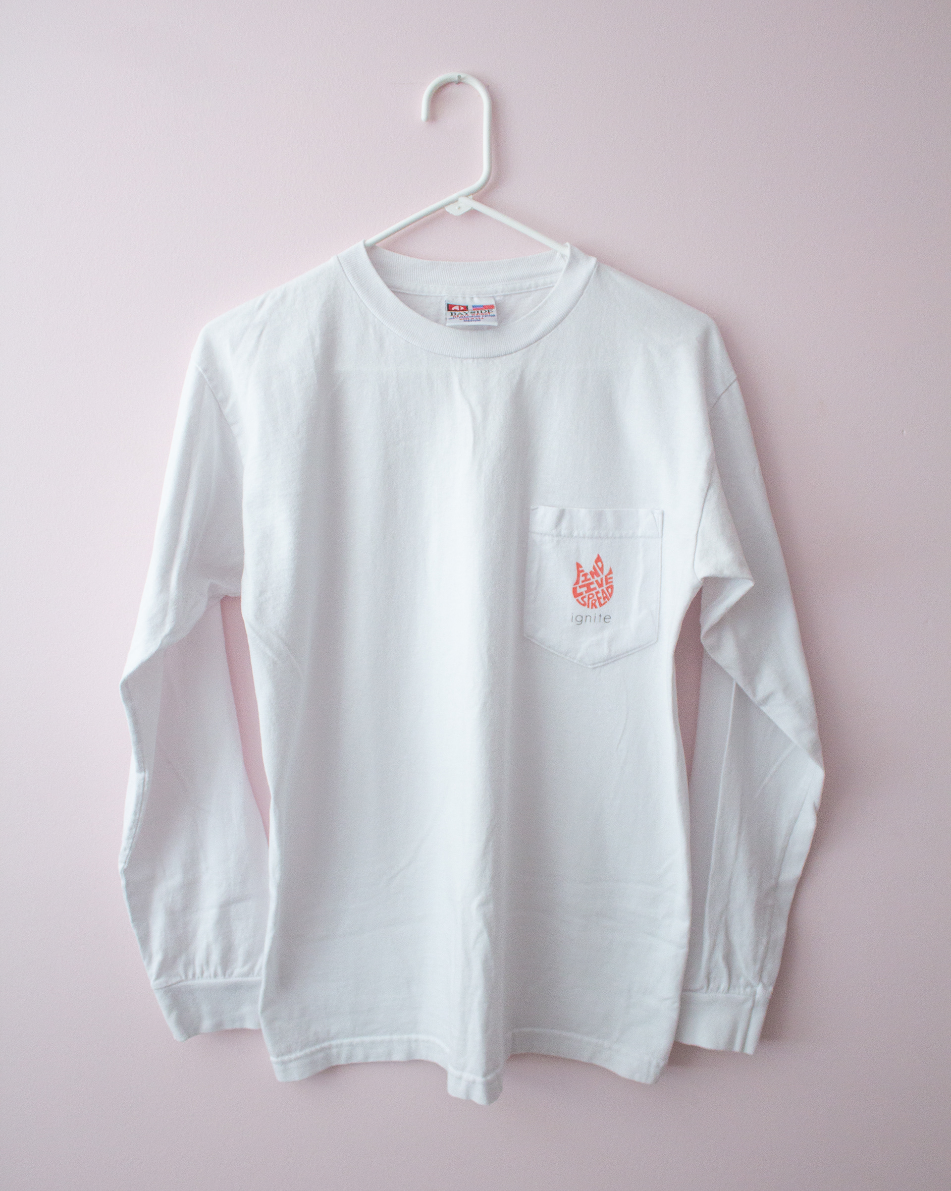 A white long sleeve pocket shirt with an orange flame logo made of the words "FIND", "LIVE", "SPREAD". Below the logo is the word "ignite" in thin black letters.