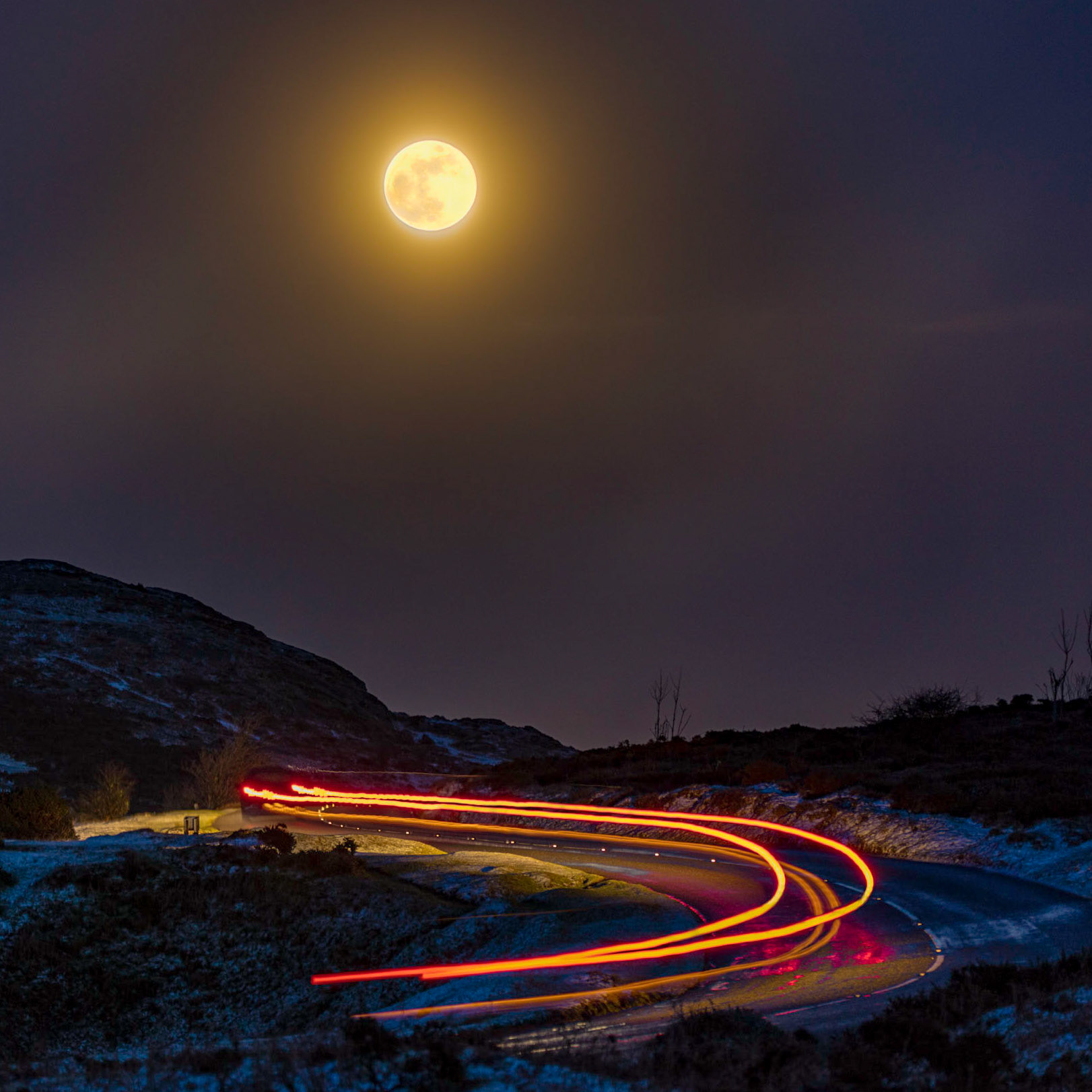 Elongated tail lights of a passing car under a Super/Blue Moon rising over Saddle Tor in Dartmoor National Park.