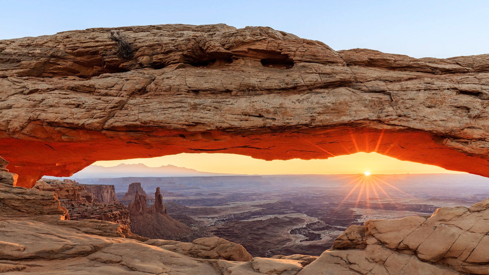 The sun breaking over the horizon and illuminating the underside of Mesa Arch perched on the side of a cliff overlooking the canyon in the Island of the Sky section of Canyonlands National Park, Utah, USA.