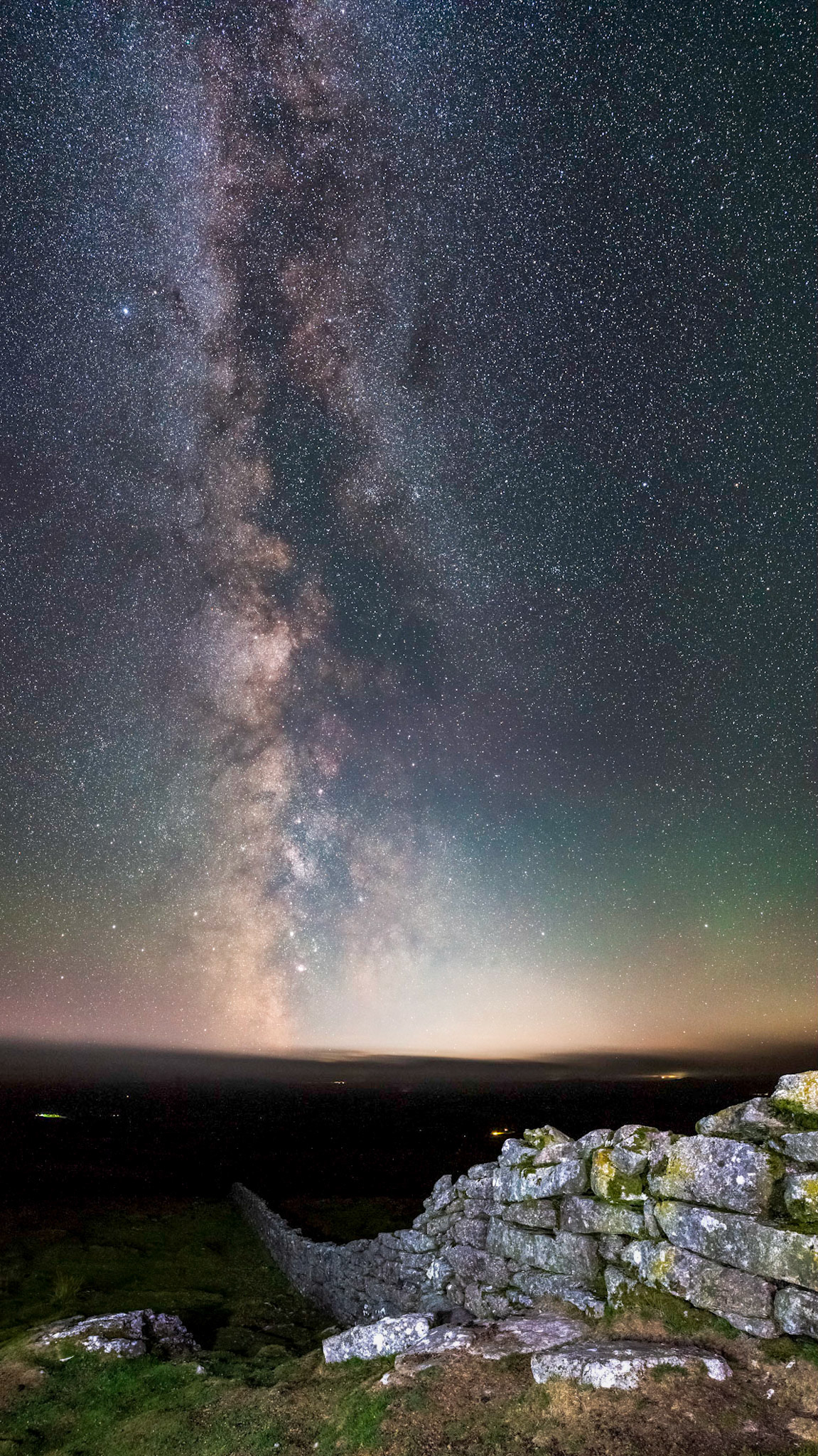 An old stone stacked wall extending down the hill and into darkness as the Milky Way extends into the sky in Dartmoor National Park near Hameldown.