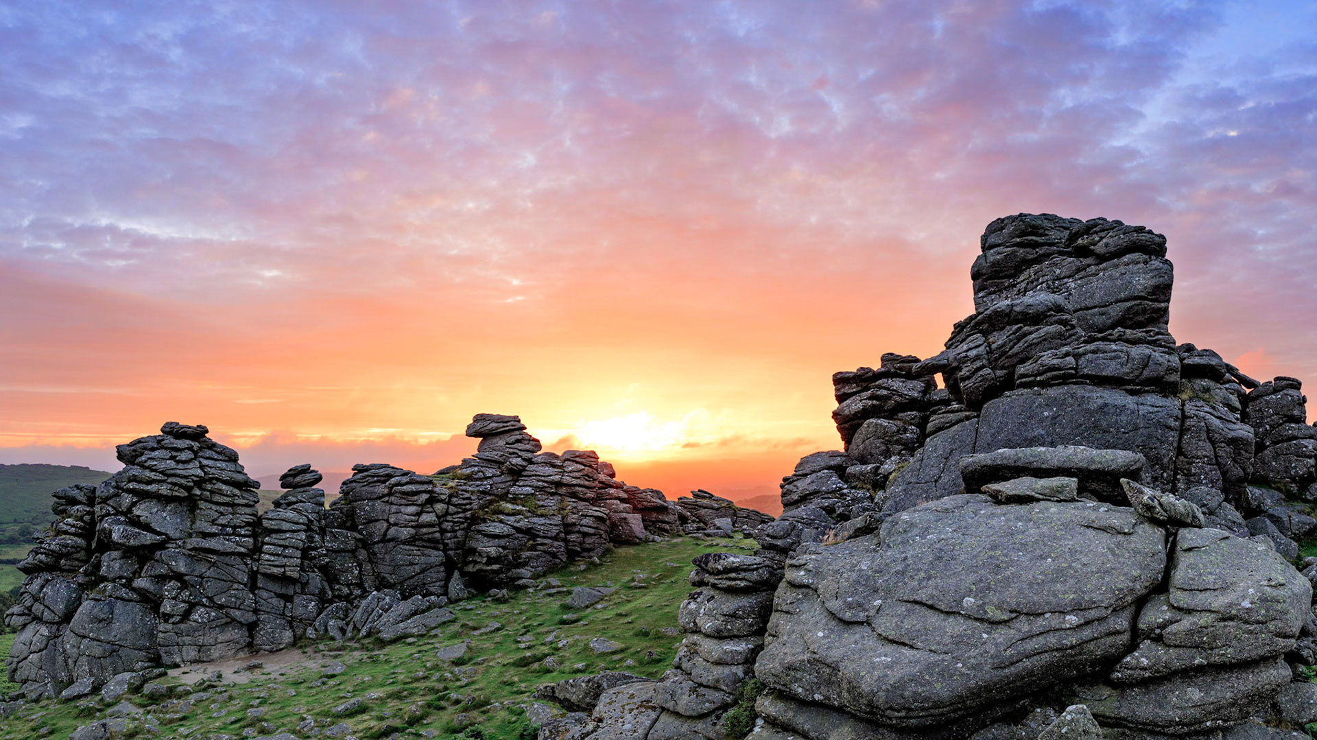 The morning sun shining through breaks in the clouds to illuminate the sky on Hounds Tor in Dartmoor National Park.