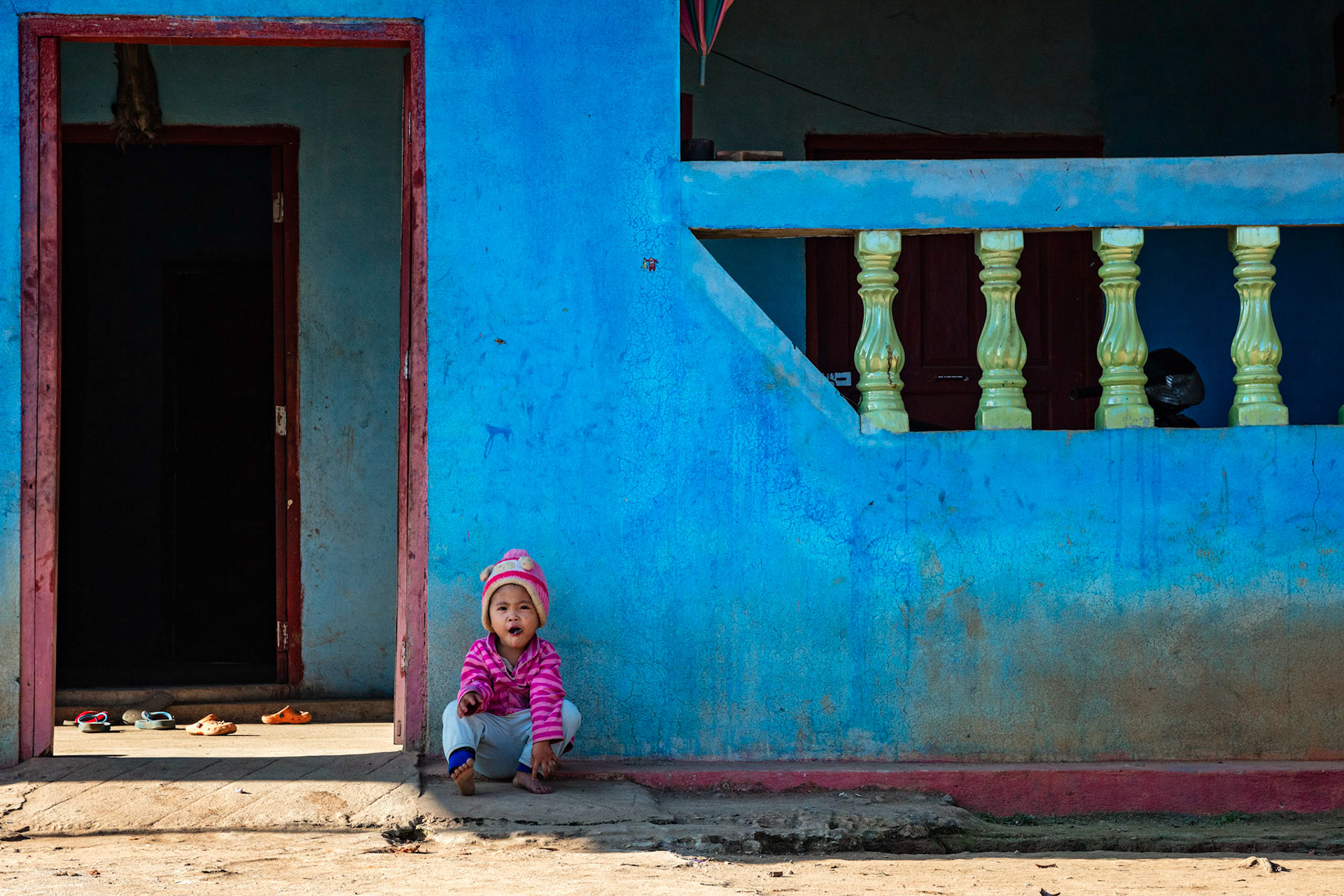 Little girl sitting outside of her home wearing colorful clothing backed by a color wall.