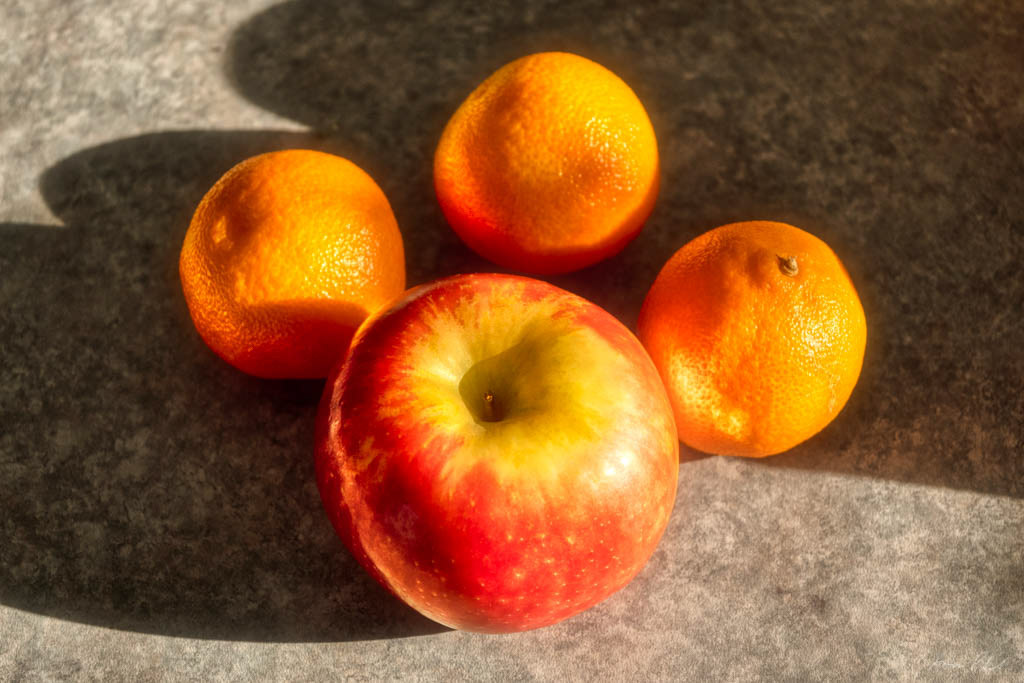 close up photo of three oranges and an apple in early morning sunlight