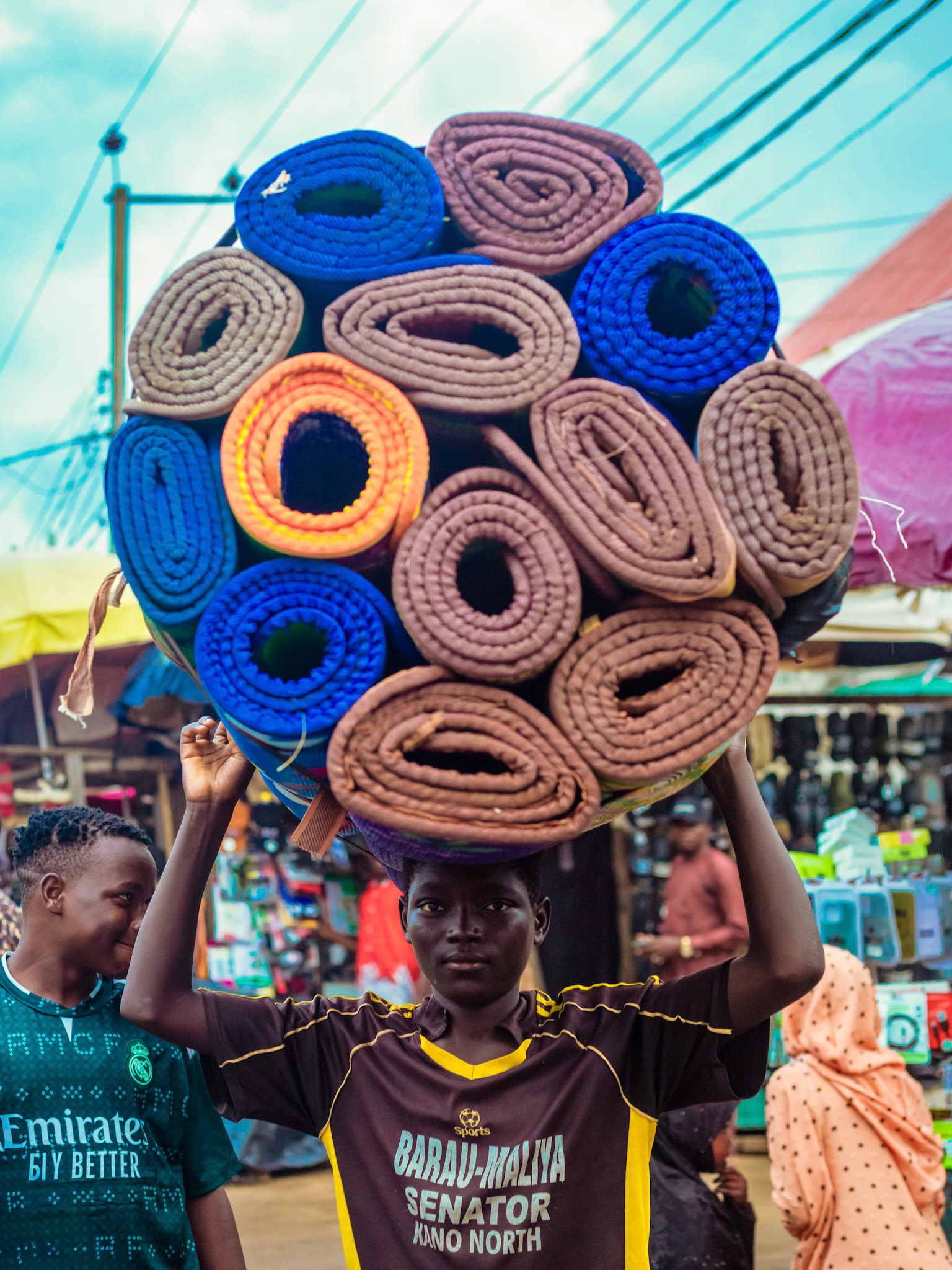 A young vendor carries a towering arrangement of rolled mats through a busy market, creating a striking study of labour, balance, colour and economic survival.