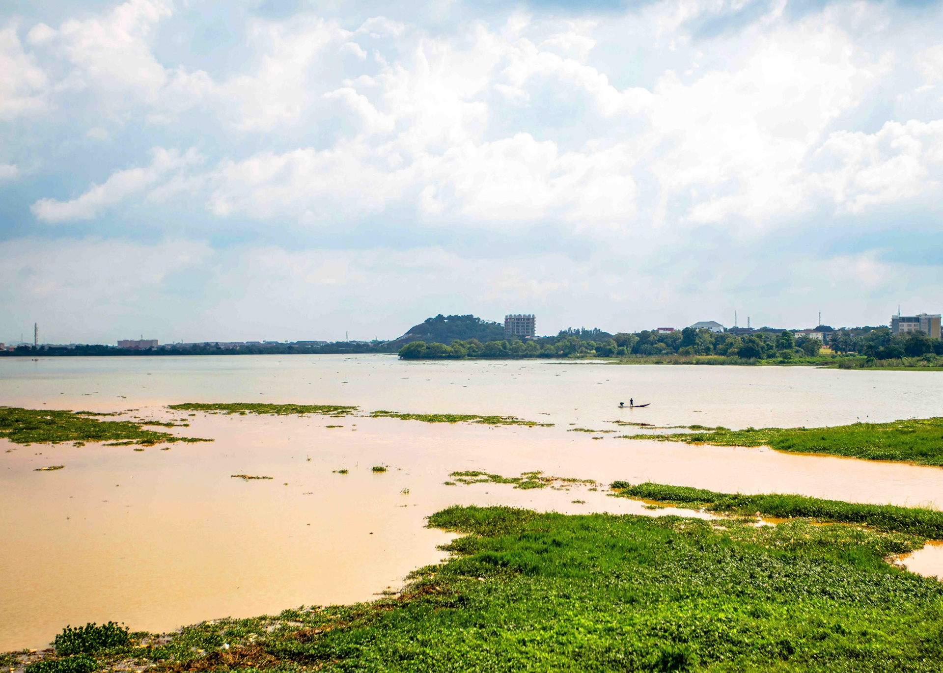 A wide expanse of water meets a distant landscape where nature and human life intersect. A small boat and floating vegetation create a quiet reflection on movement, memory and human presence.
