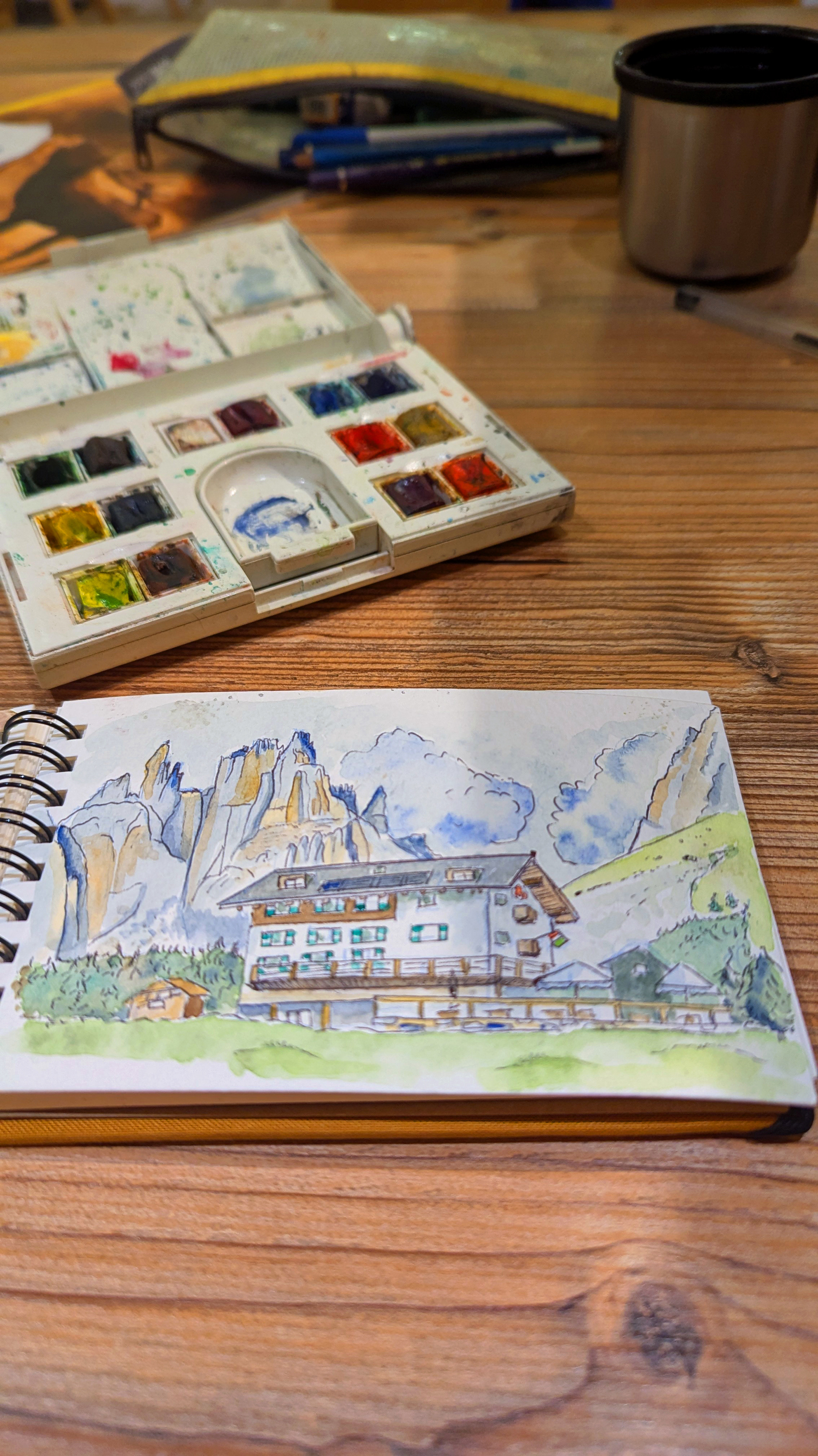 Photo d'une page de min carnet de croquis représentant un refuge dans les dolomites en italie, avec mon aquarelle et ma trousse ouverte en arrière plan 