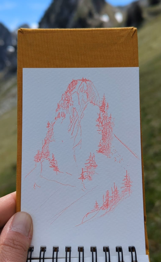 Présentation d'un dessin au crayon de couleur rouge dessiner sur un carnet de croquis en pleine montagne. On y cvoit un sommet avec des sapins. Dans le massif des Bauges