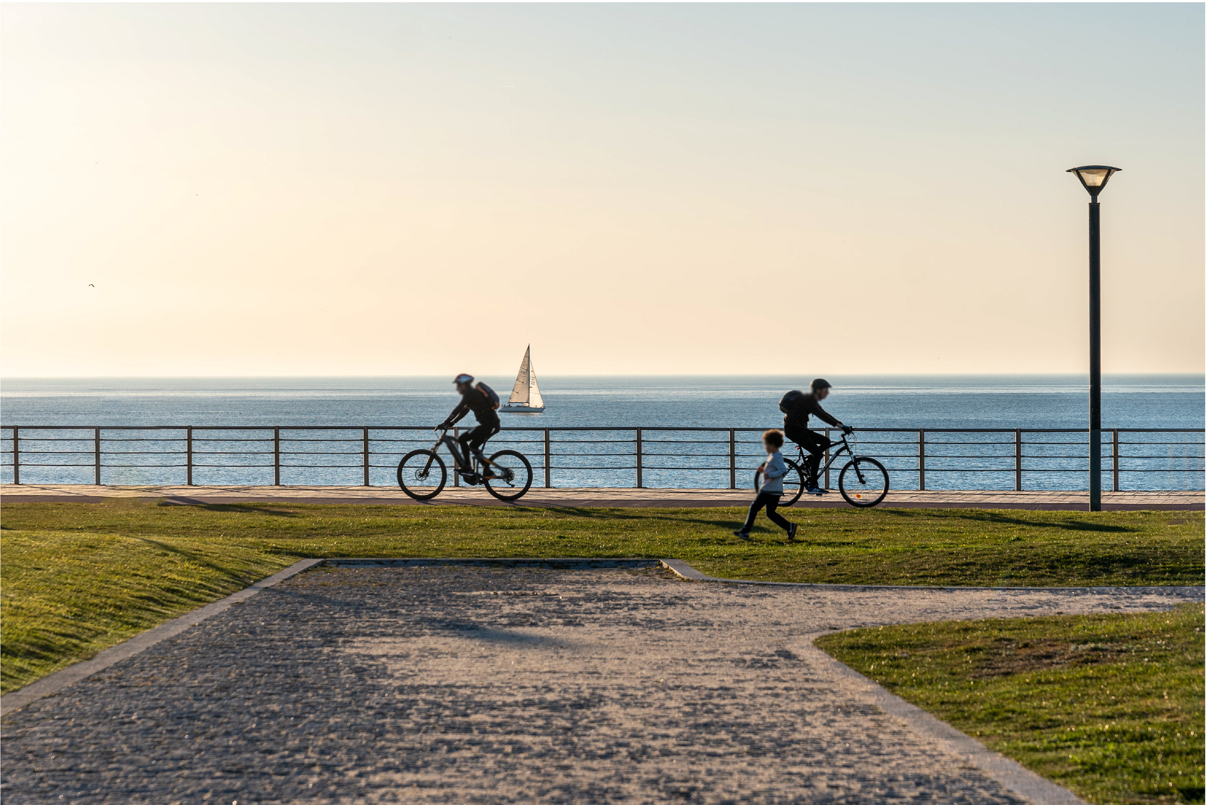 Fotografia do passeio da praia de Valadores, Gaia, com dois ciclistas, uma criança a correr e um veleiro ao fundo.