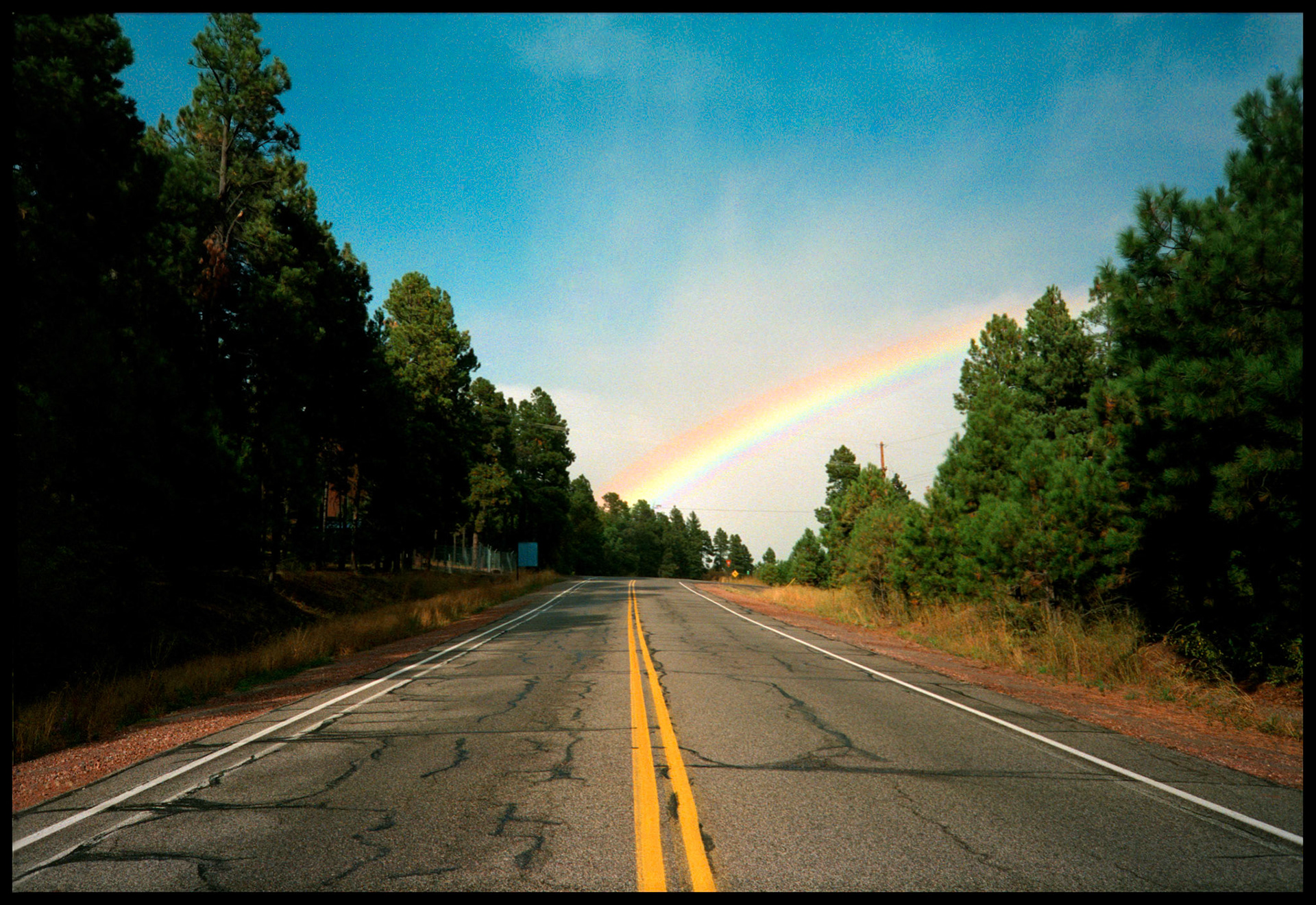 The double yellow lines of a lonely Highway in New Mexico seemingly leading to the base of a rainbow; evoking idealism, optimism, and a sense of a reward at the end of your journey. Near Los Alamos New Mexico. 1993