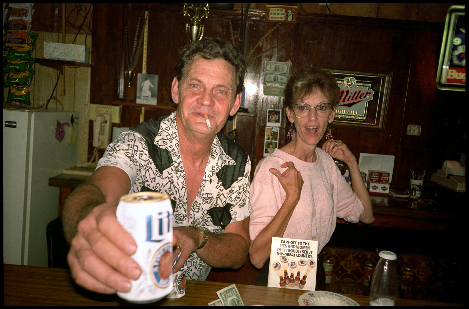 A small town tavern owner and his wife behind the bar with the owner greeting the photographer by offering him a beer.  Greencastle Missouri 1991